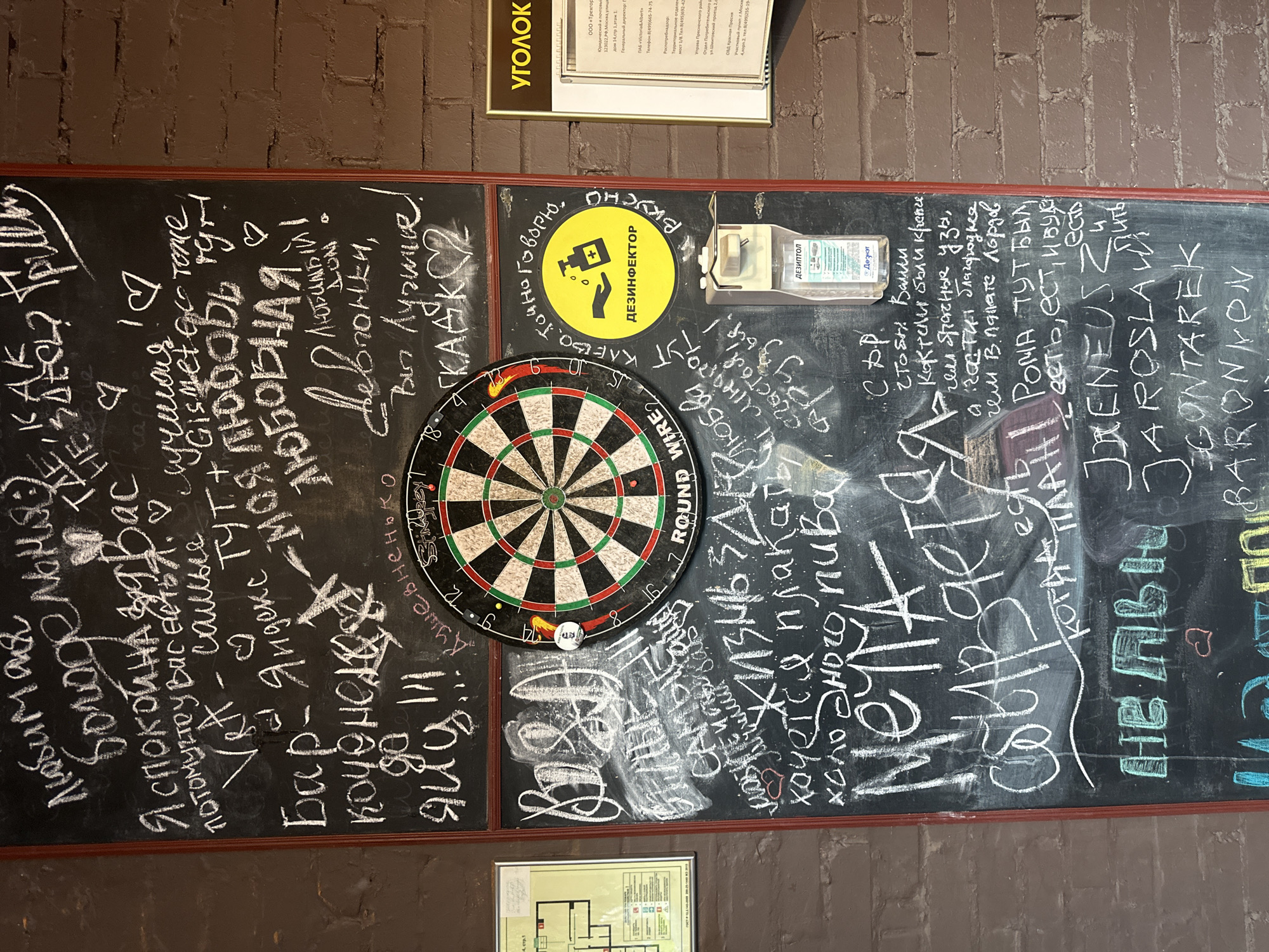 The image displays a black chalkboard adorned with various messages, names, and a dartboard, set against a brick wall within what appears to be a casual establishment in Moscow, Russia. **Subjects and Objects:** * **Chalkboard:** The primary subject, covered in white chalk text and drawings. It measures approximately 1.8 meters tall by 1 meter wide. * **Dartboard:** A standard dartboard, marked "ROUND WIRE," is centrally mounted on the upper half of the chalkboard. A single red-fletched dart is stuck in the "20" segment. * **Hand Sanitizer Dispenser:** A white, wall-mounted liquid soap/sanitizer dispenser is affixed to the chalkboard below the dartboard. Its clear reservoir shows a blue liquid, and a label reads "ДЕЗИТОЛ" (DEZITOL). * **Signs:** * To the right of the chalkboard, on a reddish-brown brick wall, is a yellow-bordered sign with the word "УГОЛОК" (CORNER) at the top. Below it, a regulatory sign provides an address: "123022, РФ, Москва, улица 190... дом 14, стр. 1, этаж 1," confirming the Moscow location. * To the left, on a dark brown wall, a light-colored sign displays a floor plan or schematic, mostly unreadable. * **Walls:** The surrounding walls are constructed of reddish-brown bricks on the right and a dark, possibly painted, surface on the left. **Chalkboard Text and Drawings (translated from Russian):** The text is a collection of informal messages, expressions, and names: * **Top Left:** "Любимая Богодельня" (Beloved Almshouse/Charity house – used affectionately for a simple, welcoming place). * **Upper Left:** "Я спокойна за Вас потому что у Вас есть: Ver - самые лучшие" (I am calm for you because you have: Ver - the very best ones). * **Upper Middle:** "КАК НЕ ЗДЕСЬ?" (How not here?). "Бар - Яндекс ТУТ + Gismet go токе" (Bar - Yandex IS HERE + Gismet go token/talk – referencing Yandex and Gismeteo, a weather service). * **Upper Right:** "Я любовь / ЛЮБОВНАЯ" (I am Love / LOVING). "девочки, Вы лучшие!" (Girls, you are the best!). "СКАДК" (SKADK – likely an initialism or nickname). * **Middle Left:** "Коченелая до яиц!!!" (Frozen to the balls!!! – a vulgar expression for being extremely cold). * **Around the dartboard (yellow circle):** "КЛЕВО, ТОЧНО ГОВОРЮ, ВКУСНО" (Cool, I'm telling you, tasty). Inside the yellow circle, an icon of hands under a dispenser with a cross symbol, and the word "ДЕЗИНФЕКТОР" (DISINFECTOR). * **Below dartboard, left:** "ВЖОД!!!" (Unclear exclamation). "САМЫЕ САМЫЕ ЛЮБИМЫЕ" (The very very beloved ones). *
