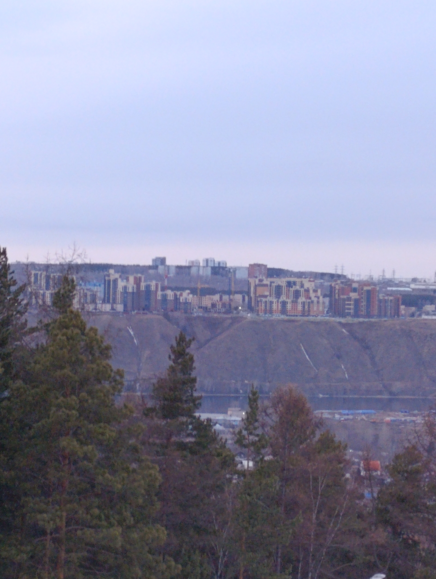The image presents a cityscape from an elevated perspective, most likely in the city of Krasnoyarsk, Russia. The foreground consists of trees, partially obscuring the view. In the midground, a long, elevated landform supports a developed urban area with numerous multi-story buildings and infrastructure. The background is filled with a pale sky. A river or large water body is faintly visible between the elevated landform and foreground trees. No people are visible.