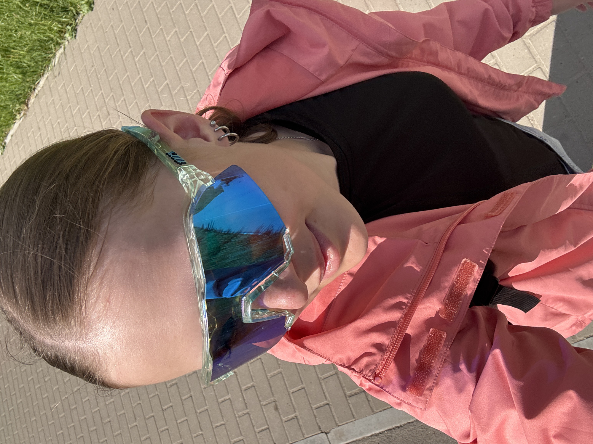 A woman with blond hair, fair skin, and piercings in her left ear is wearing blue, angular sunglasses and a pink jacket over a black top. She is outdoors on a brick walkway, with grass visible on the left side of the frame. The location is likely in the city of Chaikovsky, Russia.