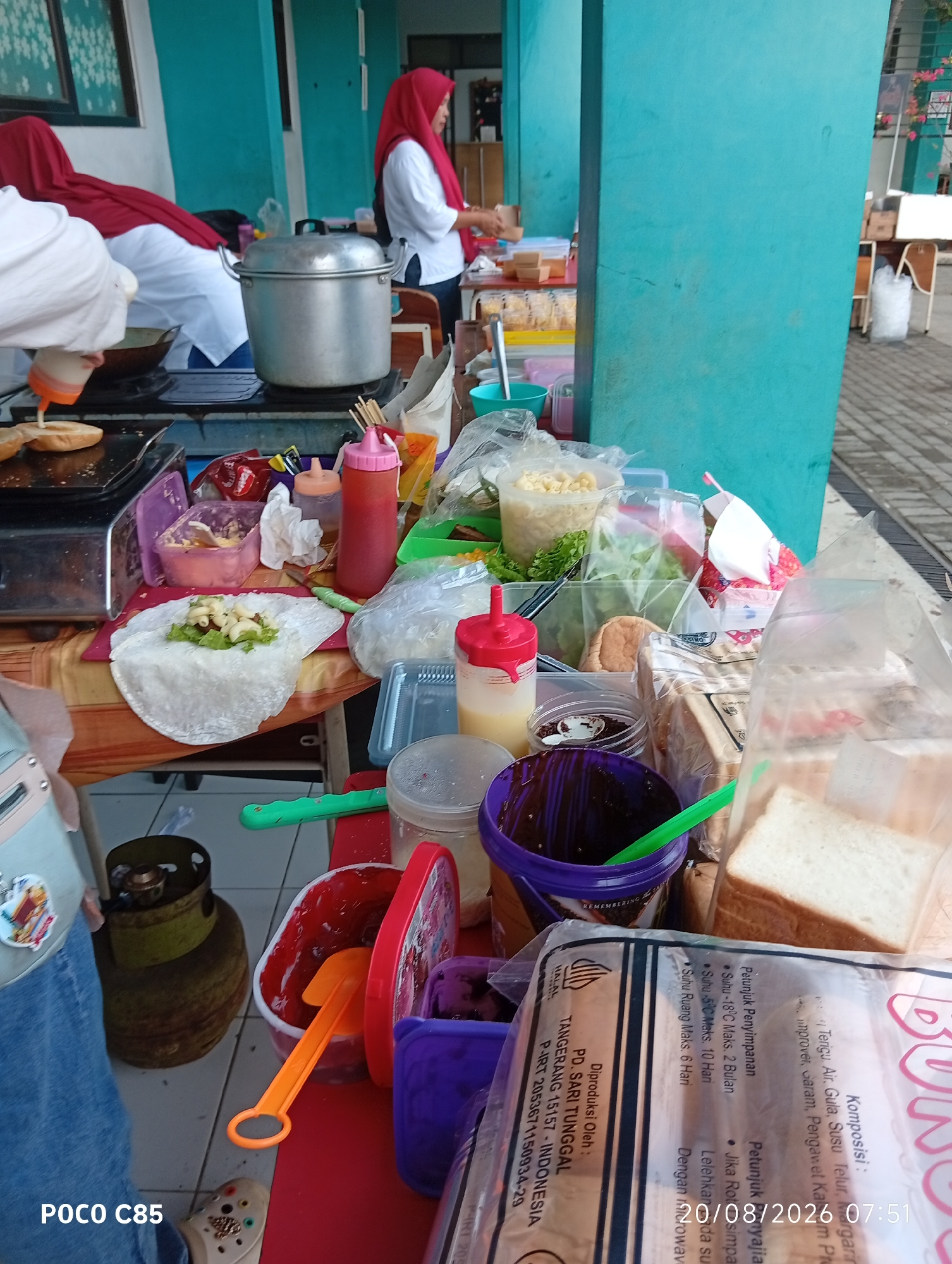 The image depicts an outdoor food stall in South Tangerang, Indonesia, likely during daylight hours given the lighting. A person wearing a red hijab and a white shirt is visible in the background, appearing to be preparing food. In the foreground, a variety of food items and ingredients are arranged on a table, suggesting a street food business. There are containers of sauces, lettuce, what appear to be slices of bread in plastic packaging, and a large pot. A green propane tank is also visible on the ground next to the table. The overall scene suggests a busy food preparation area, possibly at a school or public event, given the casual setup and the presence of multiple people in the background. The text "POCO C85" is watermarked on the bottom left, indicating the device used for capture. The date stamp "20/08/2026 07:51" also appears in the image.