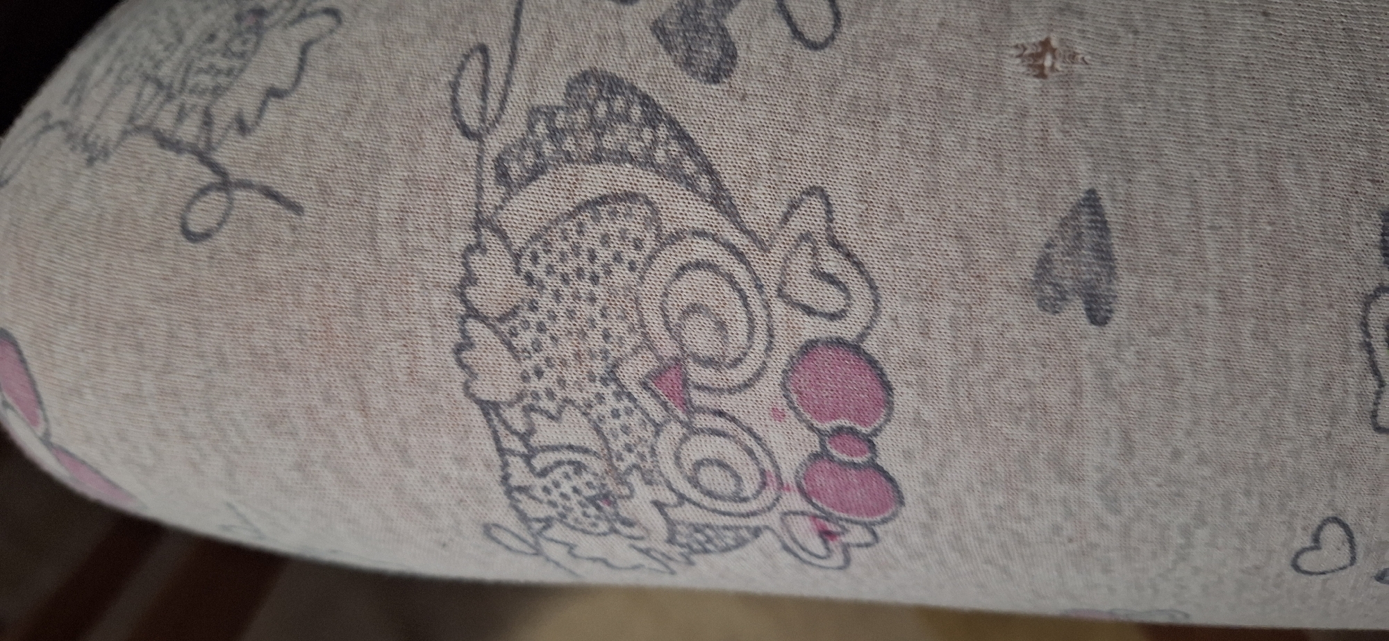 The image shows a close-up of a patterned fabric, likely covering a bolster or pillow. The fabric is a light beige color with printed designs in grey and pink. The designs include depictions of what appears to be a cartoonish stylized face, possibly a cat-like character with a large head, swirls, and a pink bow. There are also smaller grey heart shapes and what seems to be outlines of other figures in the background. A small hole is visible in the fabric near the top right. The environment appears to be indoors, with the base of the bolster or pillow resting on a dark-colored surface. There is no information to assess the location in the city of Blagoevgrad, Bulgaria.