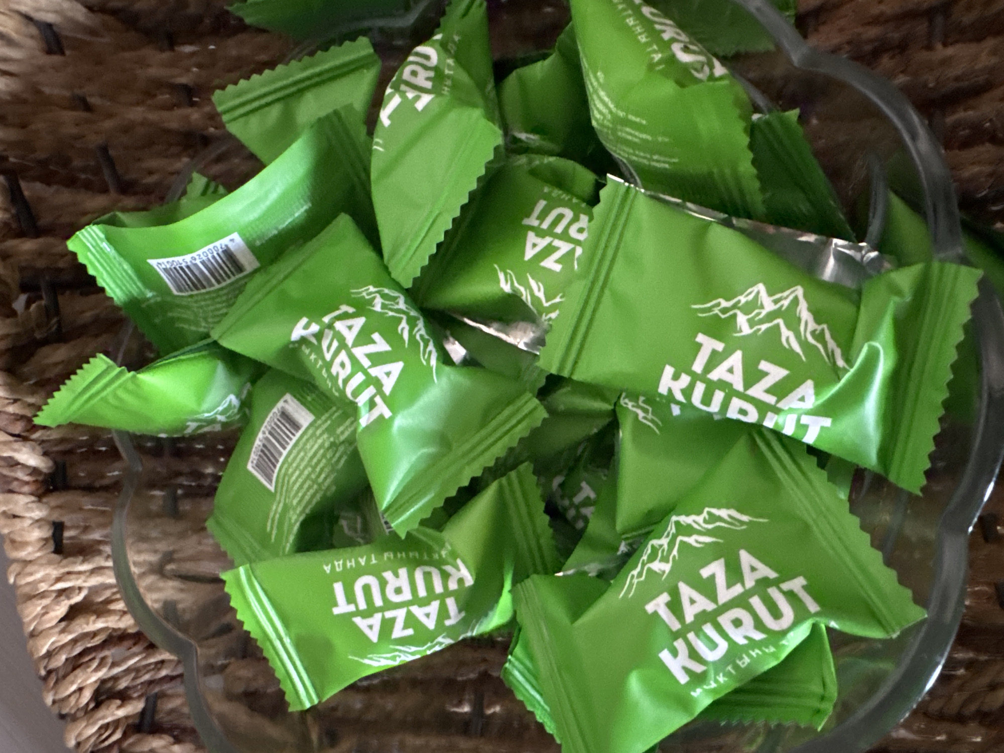 The image displays a close-up, overhead view of a glass bowl filled with individually wrapped candies. The candies are all wrapped in bright green foil packaging with white lettering. The text on the packaging appears to say "TAZA KURUT" in large, bold letters. There are also some smaller words in what looks like Cyrillic script. Some of the wrappers have images of mountains. The bowl is sitting on a surface that appears to be made of woven material, possibly a basket. The overall impression is of a collection of sweet treats. The image was taken in Bishkek City, Kyrgyzstan.
