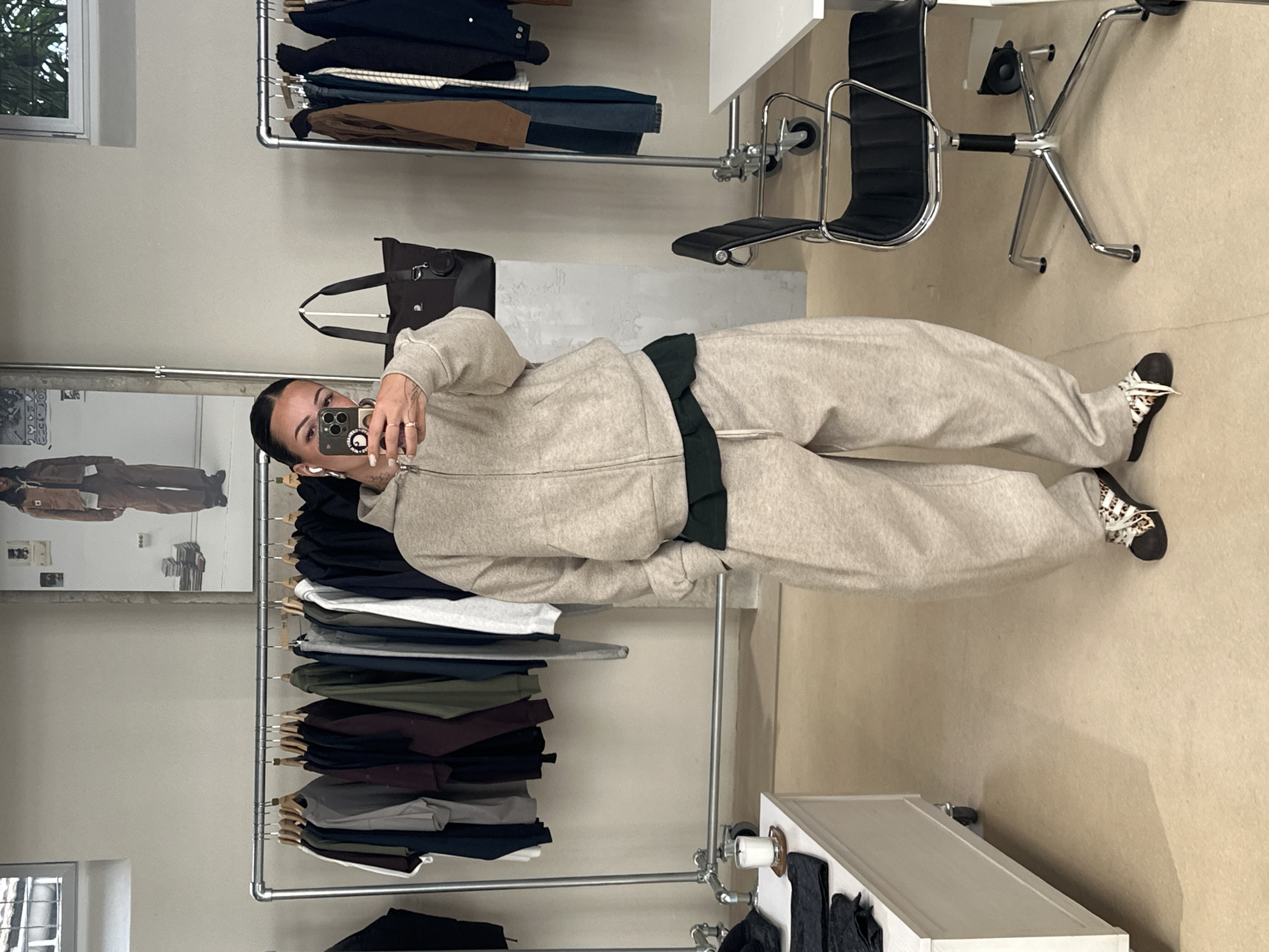A person takes a selfie in front of a mirror in a clothing store. The person is wearing a beige tracksuit and patterned sneakers. The store has racks of clothing, a desk with a chair, and a display cabinet. A reflection in the mirror suggests the presence of a window and an additional room. The image lacks sufficient detail to confirm a location in Paris, France.