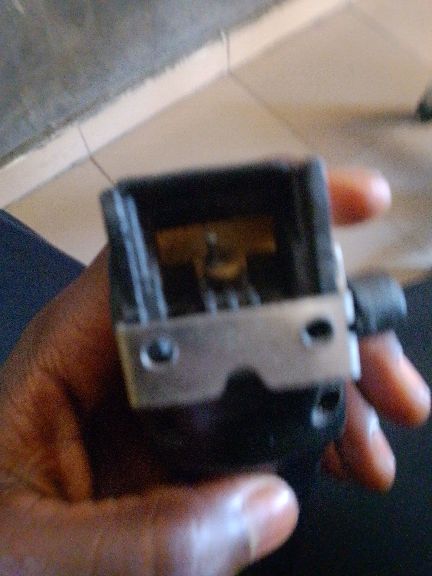 The image shows a close-up of a person's hand holding a black object with metal accents. The object appears to be some kind of electronic component or small appliance part, possibly a trimmer or a small motor housing. The hand is dark-skinned, and some fingers are visible, gripping the object. The background is blurred, showing a tiled floor and a wall, suggesting an indoor environment. The location is Jalingo, Nigeria.