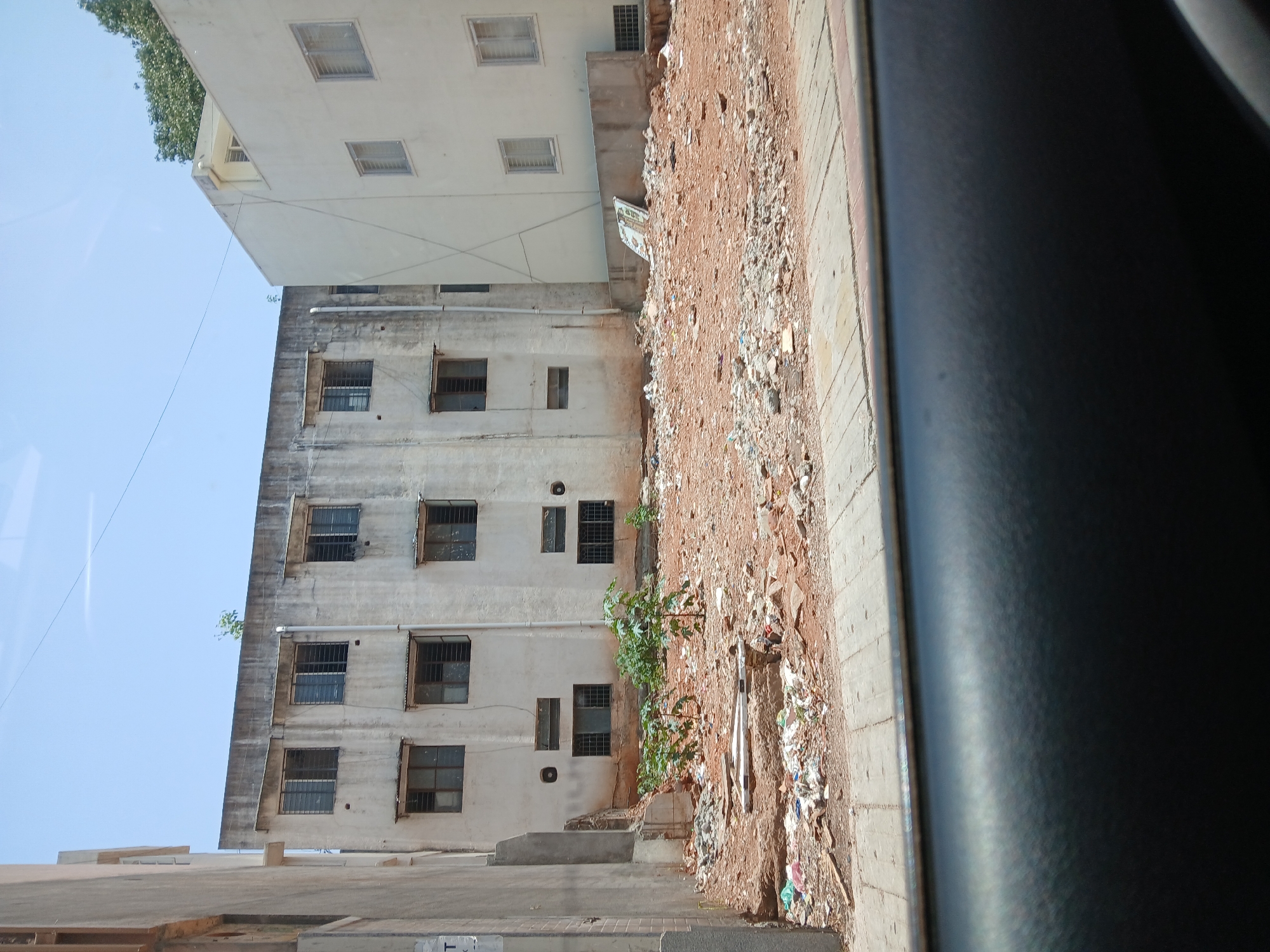 The image shows an exterior view of an apartment building taken from inside a vehicle, likely in Bengaluru, India. The building is weathered and appears to be several stories tall. The windows have metal bars. There is visible dirt, debris, and trash accumulation on the ground beside the building, bordering a concrete path or road. The sky is visible above the building, suggesting it is daytime. The window frame of the vehicle is partially visible on the right. A young tree or shrub grows between the building and the garbage heap.