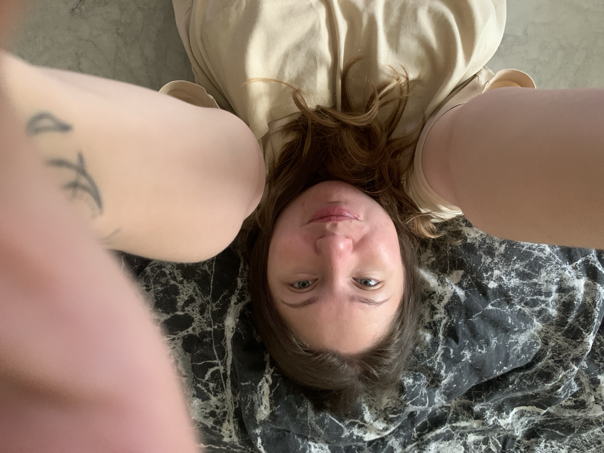A woman is lying on a black and white marble-patterned surface. She is wearing a beige jacket and has light brown hair. A tattoo is visible on her left arm. The picture is taken from an overhead perspective, with the woman's head positioned towards the bottom of the frame. No location in Chaikovsky, Russia, can be determined from the image.