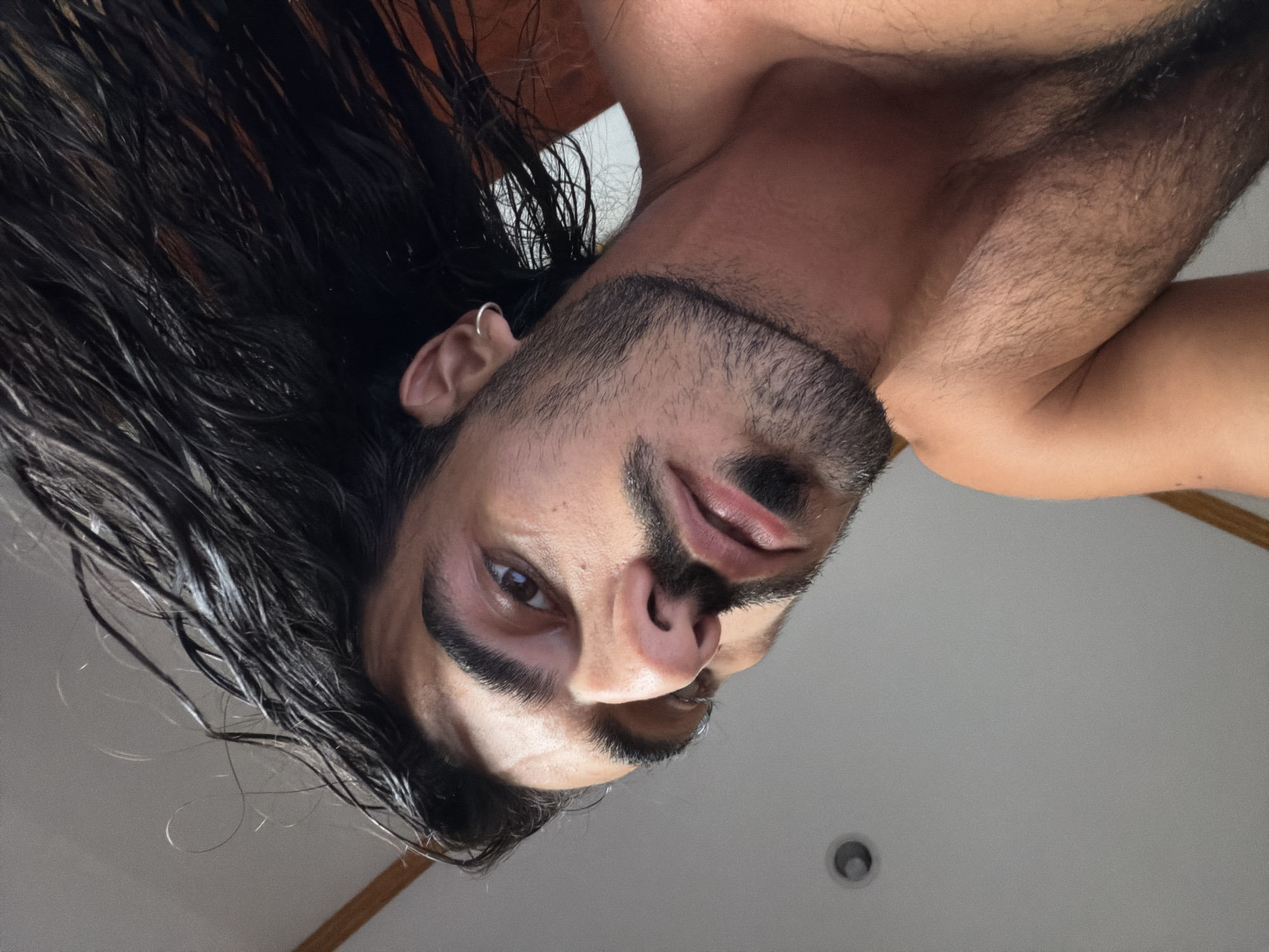 A man with dark, wet hair and a short beard is captured from a low angle, looking down at the camera. He has an earring in his left ear. His chest and shoulders are visible. The background is a plain, off-white ceiling with a recessed light fixture. The image was taken from an unknown location in North Kuta, Indonesia.