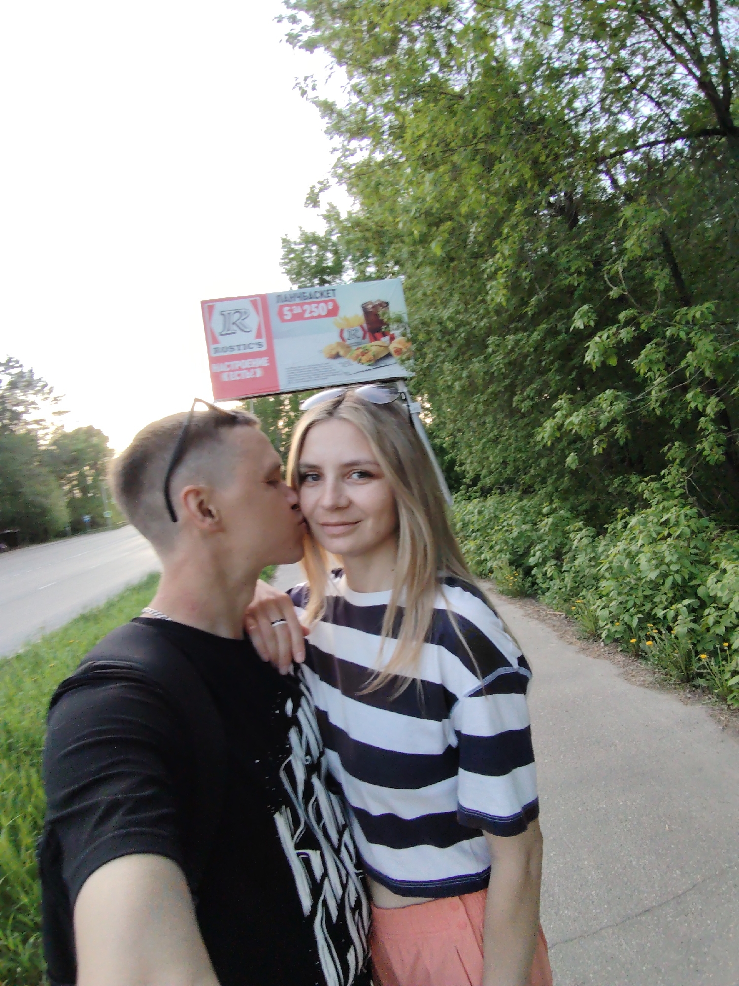 The image shows a couple on a paved path next to a road in what appears to be a park or green space. The background includes trees and other vegetation. An advertising billboard is visible in the background. The man is kissing the woman on the cheek. The location is Chaikovsky, Russia.