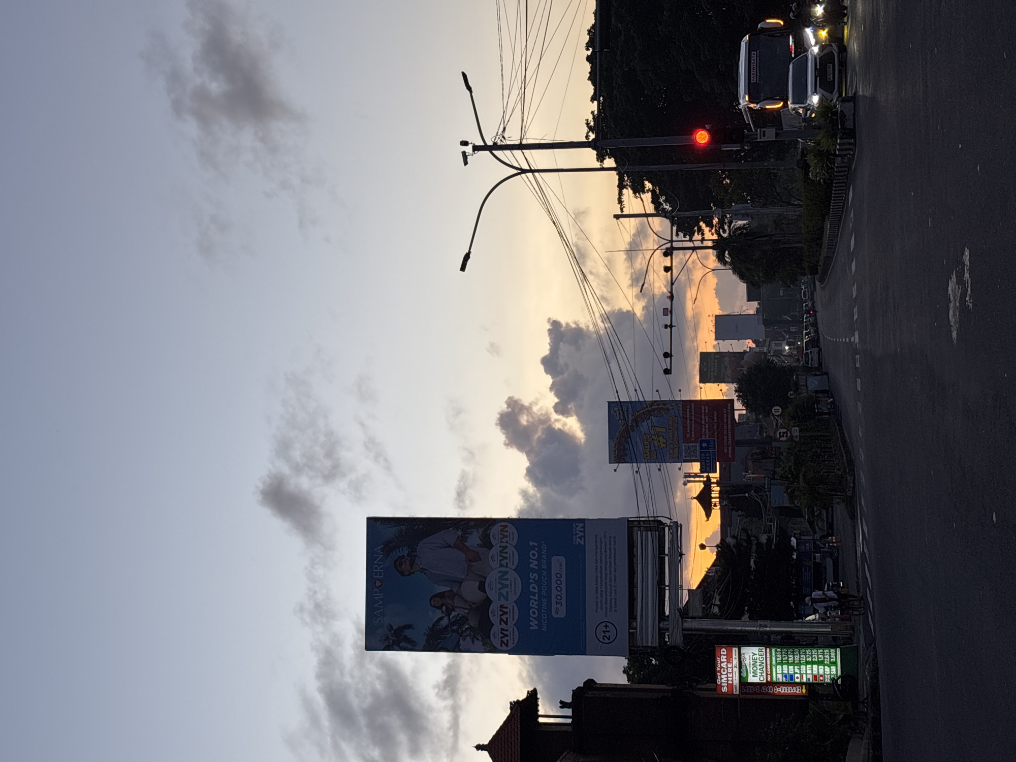 The image captures a street scene at dusk in Kuta, Indonesia. A two-lane road stretches into the distance, flanked by buildings and trees. On the right side of the road, a bus and a white car are visible, waiting at a red traffic light. Utility poles and wires crisscross the sky. Two prominent billboards stand on the left, advertising "ZYN" nicotine pouches and Balis. Below the nicotine billboard is a "Money Changer" sign, displaying exchange rates. The sky is a mix of gray clouds and orange hues from the setting sun. The overall lighting is subdued, with some areas appearing dark. There are additional billboards in the mid-distance, and some vehicles traveling the road into the distance.
