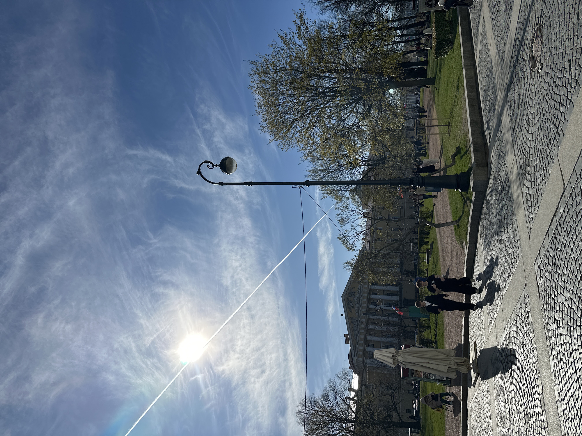 This outdoor image captures a sunny day in Saint Petersburg, Russia, showcasing a blend of urban architecture and park greenery. The photo appears to be taken with a vertical orientation, making the sky dominate the left side and the ground the right.
The sky is a vibrant blue, adorned with wispy cirrus clouds. A bright, low sun causes a distinct lens flare near the bottom left, and a long, white airplane contrail slices diagonally across the entire sky. A classic, ornate street lamp with a spherical light fixture stands prominently in the mid-ground.
On the ground level, a wide paved area with intricate cobblestone patterns stretches across the right. Adjacent to it is a well-maintained grassy park area dotted with trees, some just beginning to show new spring foliage, while others remain bare. Long, dark shadows are cast across the paved ground, indicating either early morning or late afternoon.
Several individuals are present in the scene. Two figures, possibly men dressed in dark suits or uniforms, walk on the paved path towards the foreground. Their elongated shadows stretch behind them. Further into the background, other indistinct people are scattered throughout the park. A large, light-colored umbrella, possibly belonging to a cafe or vendor, is visible on the paved area.
In the distant background, a grand classical building with columns and decorative elements under its roofline stands against the sky, a characteristic architectural style of Saint Petersburg. The overall atmosphere is bright and calm, with people enjoying the pleasant weather. No discernible text is visible in the image.