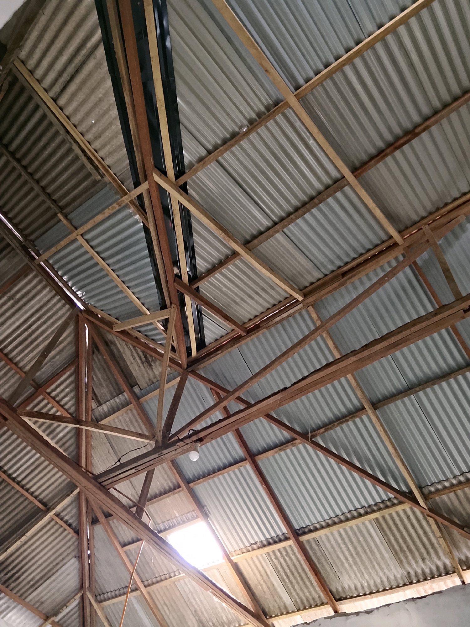 The image shows the underside of a corrugated metal roof supported by a wooden beam structure. The roof appears to be under construction or in a state of disrepair. Several wooden beams of varying sizes and conditions are visible, forming a complex framework. Some beams are weathered and appear to be repurposed. The corrugated metal sheets are arranged in parallel rows, and in some areas, gaps between the sheets allow light to filter through, creating illuminated patches. A single light bulb hangs from a wire attached to one of the beams. The overall scene suggests an unfinished or rustic building located in Karang Gading, Indonesia.