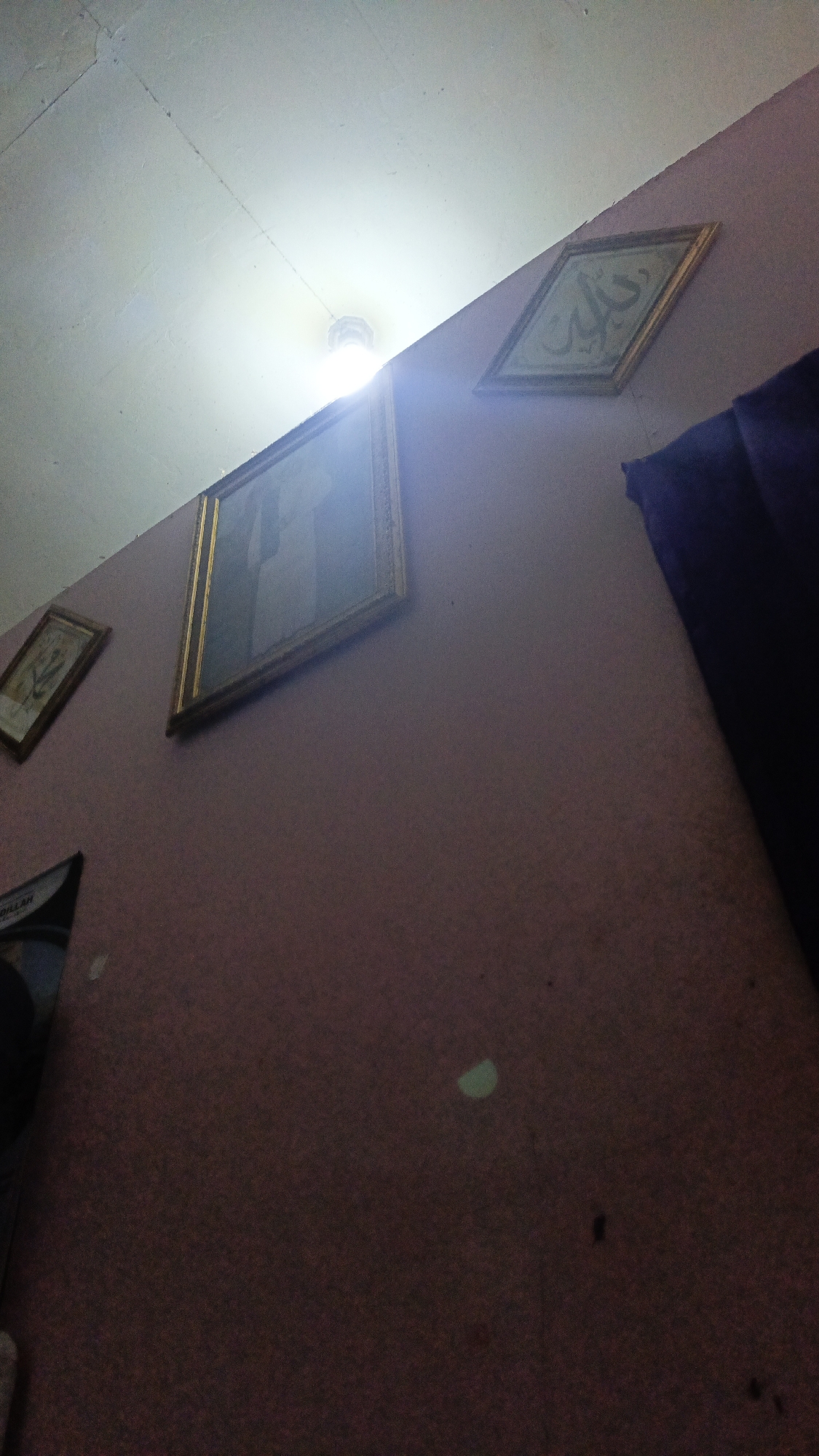 The image shows an interior wall with three framed pictures hanging on it. The wall is painted a muted pinkish-brown color. A bright light source, possibly a ceiling light, is visible at the top center of the image, casting a glare and illuminating the ceiling which appears to be white and textured.
The leftmost picture is small and rectangular, with a gold frame. It contains an image that is difficult to discern due to lighting and resolution but appears to be a figure or scene.
The middle picture is larger and rectangular, also with a gold frame. The glass front reflects the light and the image within is a portrait of a person in traditional attire, possibly religious. The face is not clearly visible due to the reflection.
The rightmost picture is framed in gold and is diamond-shaped. It contains Arabic calligraphy in a light color against a darker background.
To the right of the diamond-shaped picture, a dark purple curtain is visible, hanging down.
The image is taken from a low angle, looking upwards towards the ceiling and the pictures on the wall. The lighting suggests a low-light environment with a strong artificial light source. The scene does not provide any specific geographical indicators to pinpoint its location within Gusunge, Indonesia, but it depicts a typical domestic or religious interior setting.