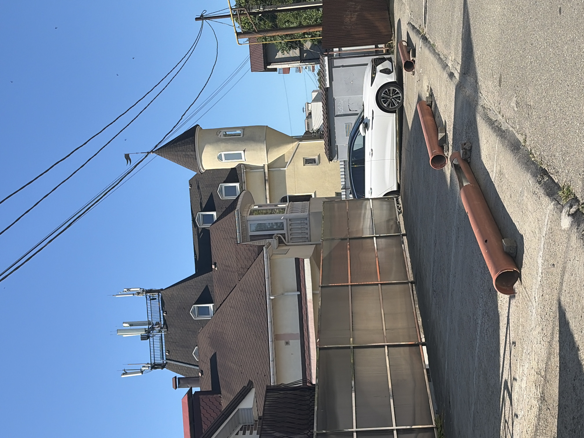 The image captures an outdoor urban or semi-urban scene in Sochi, Russia, under a bright, clear blue sky. The strong sunlight casts distinct shadows on the ground and buildings, suggesting midday or early afternoon.
On the left, a multi-story building with a dark shingled roof and several dormer windows is visible. Its rooftop hosts an extensive setup of communication antennas, likely cellular base stations, mounted on a metal structure. Adjacent to this, a distinctive building with a yellow or beige stucco facade features a prominent turret with a conical dark roof and arched windows.
A white car, possibly an SUV, is parked on a concrete surface on the right side of the frame. The foreground features a concrete pathway or driveway where several sections of terracotta-colored pipes are lying, some appearing broken or partially embedded in the ground. A translucent corrugated fence acts as a boundary between properties in the mid-ground.
Above, numerous black utility wires crisscross the expanse of the clear blue sky, supported by visible utility poles. A few small birds are seen flying high in the sky. The overall scene depicts a quiet moment in a developed area, combining residential architecture with modern telecommunications infrastructure. No discernible human activity is taking place, and no legible text is visible.