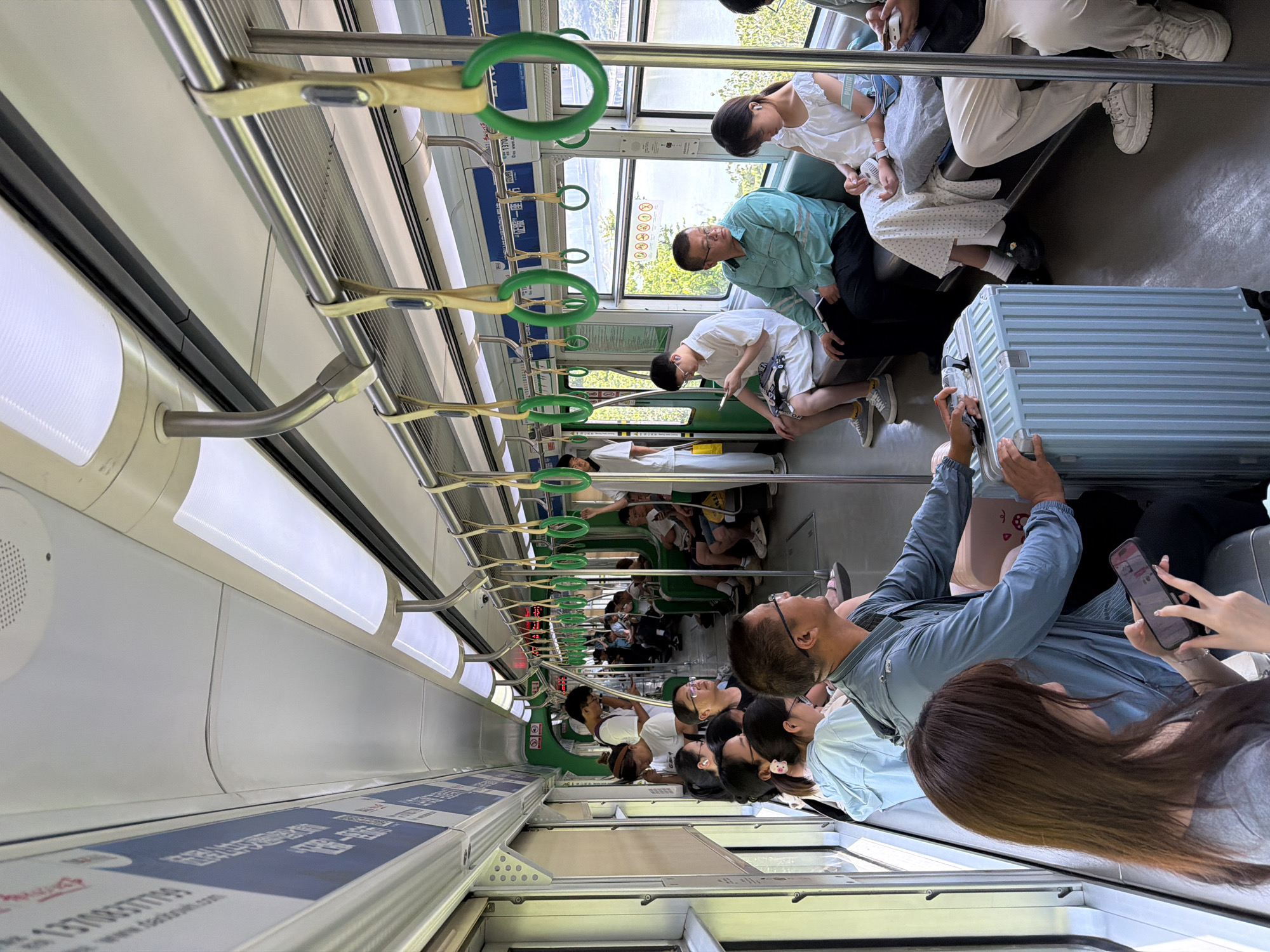 The image shows the interior of a subway train with several passengers seated and standing. The train appears to be in operation, with the scenery visible through the windows suggesting it is traveling through an urban or suburban area with greenery and a body of water. The interior of the train is brightly lit by overhead fluorescent lights and features metallic handrails and green hanging straps for passengers.
Several individuals are visible, some looking at their phones, others conversing or looking out the window. There are luggage items, including a light blue suitcase in the foreground, suggesting passengers are traveling. The people are dressed in casual summer clothing. The advertisements visible on the train's walls are in Chinese.
The scene is consistent with public transportation in Yuzhong District, Chongqing, China, given the architectural style of the train, the visible advertisements, and the surrounding landscape which often features hills and the Yangtze River in this region.