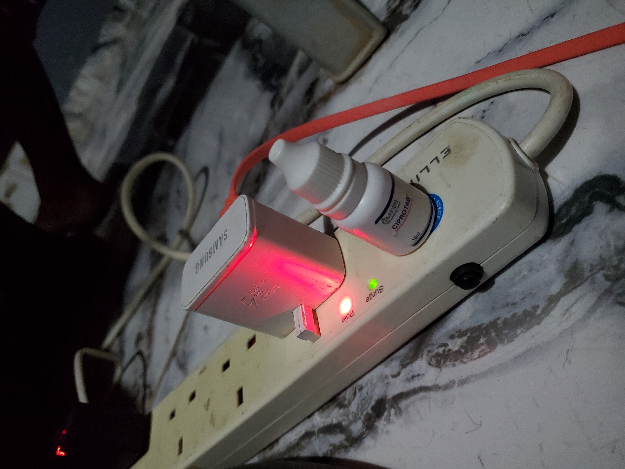 In the city of Winters, United States of America, a white multi-outlet power strip, labeled "ELLIX" (text inverted) and equipped with an illuminated green "Surge" indicator and an illuminated red "Pole" indicator (text inverted), rests on a grey-and-white mottled surface. A white Samsung "Adaptive Fast Charging" wall adapter is plugged into one of the strip's outlets, emitting a distinct red light. Adjacent to the charger, a small white dropper bottle, labeled "Caventura CIPROTAB 10ml", is positioned on the power strip. A thick beige power cable extends from the power strip, and an orange data or power cable is visible running alongside it. The background is dark and out of focus, revealing vague structural elements and additional cabling. No individuals are present within the frame.
