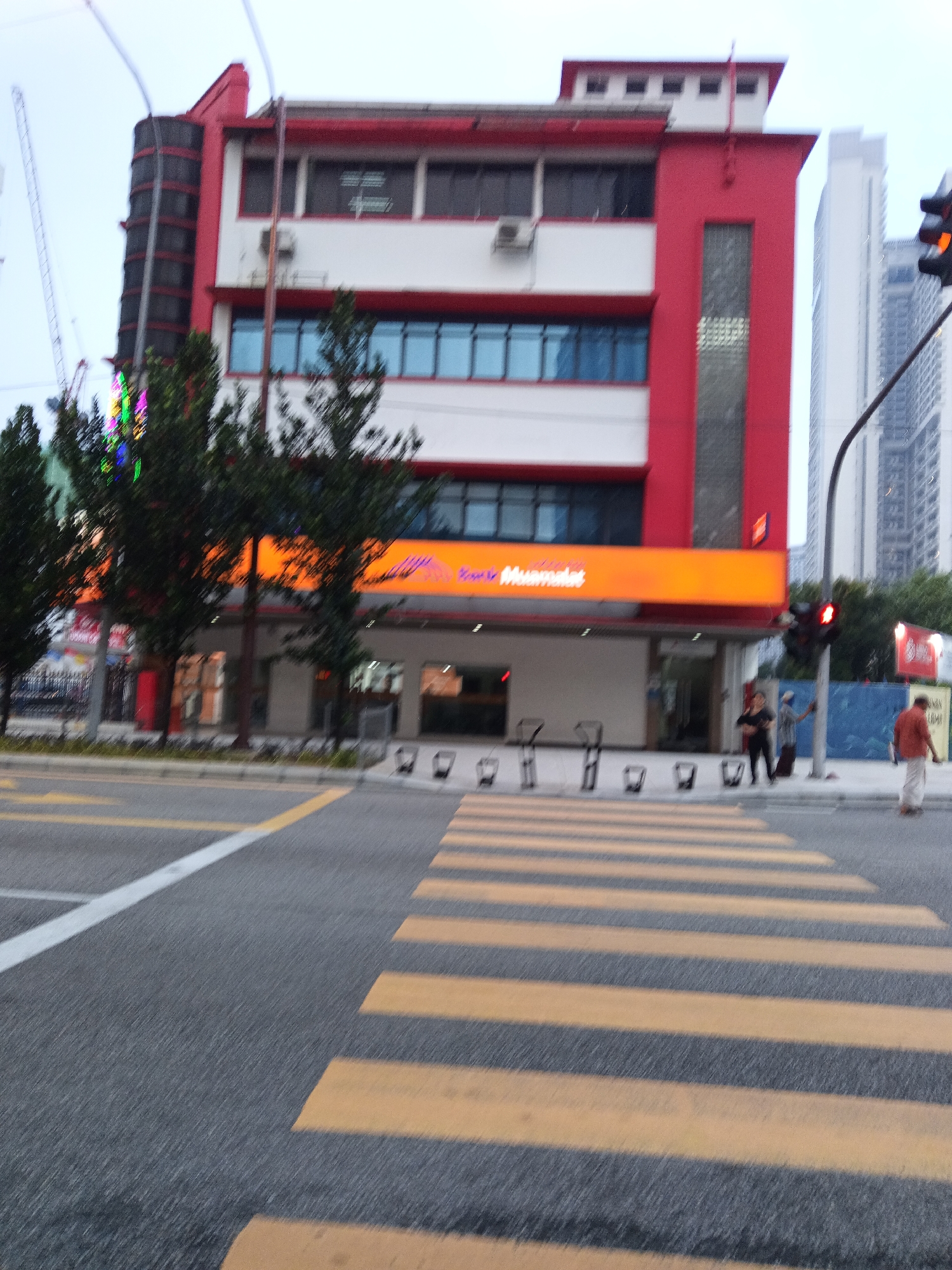 The image, taken in Kuala Lumpur, Malaysia, shows a street scene with a pedestrian crossing in the foreground. The crossing is marked with yellow and white stripes on a gray asphalt road. In the background, a red and white building with a prominent orange illuminated sign is visible. The sign appears to have text on it, possibly a business name. Trees with dark green foliage are situated in front of the building. To the right of the building, tall modern high-rise buildings can be seen in the distance. A traffic light with a red signal illuminated is positioned near the building. Several people are present, including two individuals standing on the sidewalk near the traffic light and one person walking away from the camera on the right side of the street. The sky is overcast, suggesting a cloudy day. The overall scene depicts an urban environment during daylight hours.
