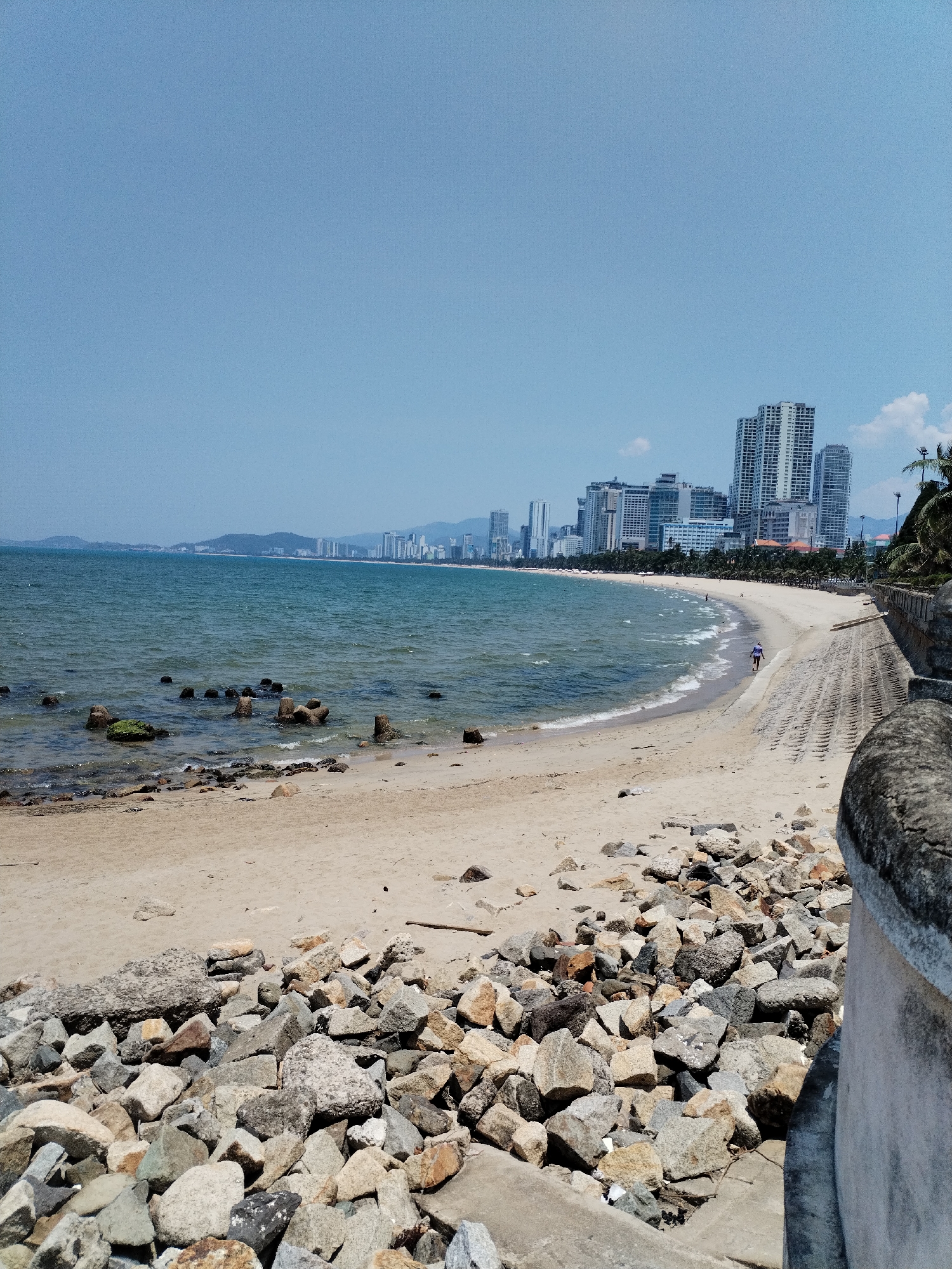 The image depicts a beach scene in Nha Trang Ward, Vietnam. The foreground consists of a pile of rocks and a concrete barrier. Beyond the rocks is a sandy beach with a few scattered objects. The ocean extends from the beach towards a cityscape in the distance. Several high-rise buildings dominate the skyline, and mountains are visible further in the background. One person is walking along the shoreline of the beach. The sky is clear and blue.
