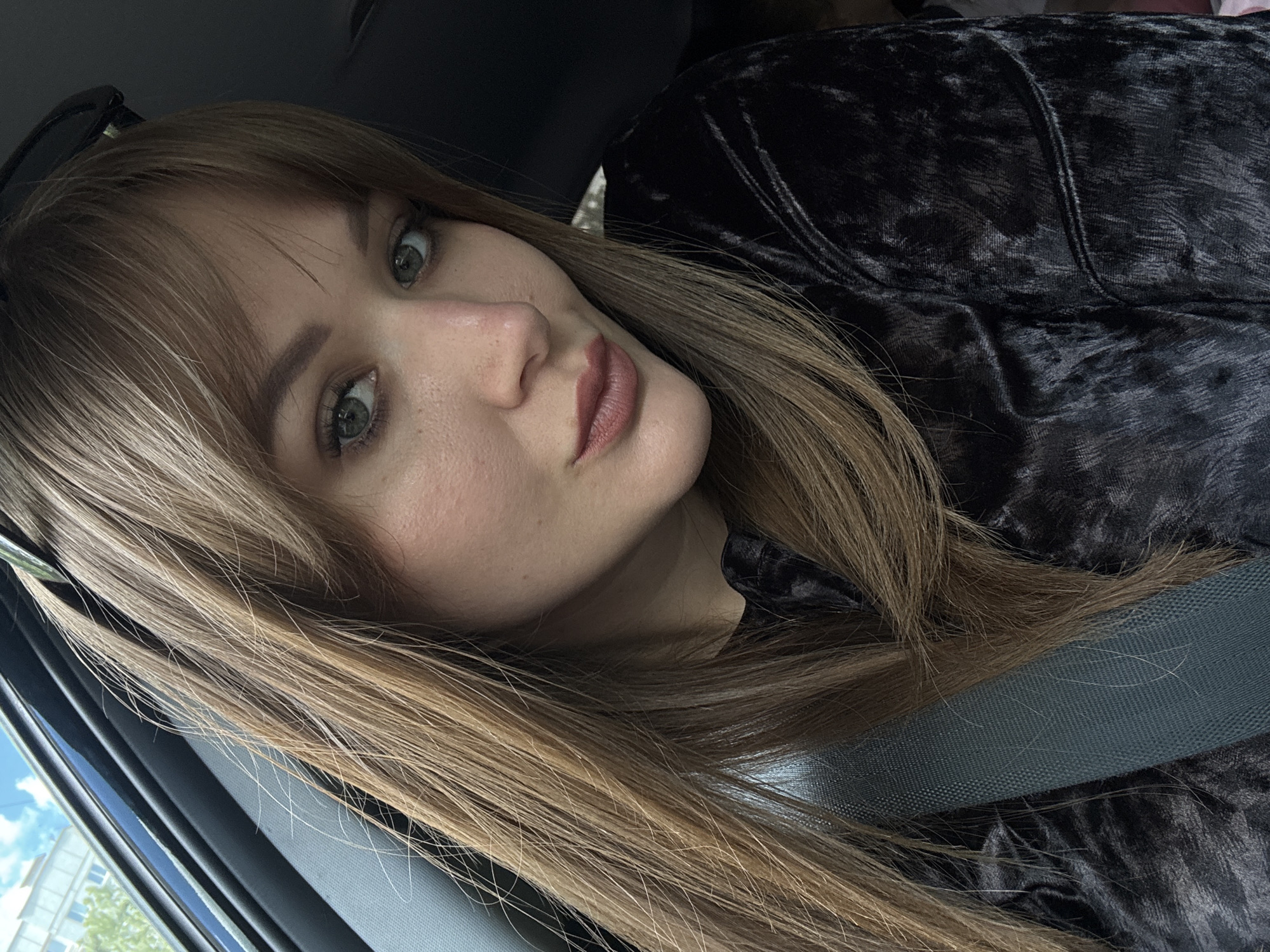 A woman with blonde hair and green eyes is in a vehicle, wearing a black velvet top and a seatbelt. The setting suggests she may be in the city of Chaikovsky, Russia, based on the presence of a building and foliage visible through the car window.