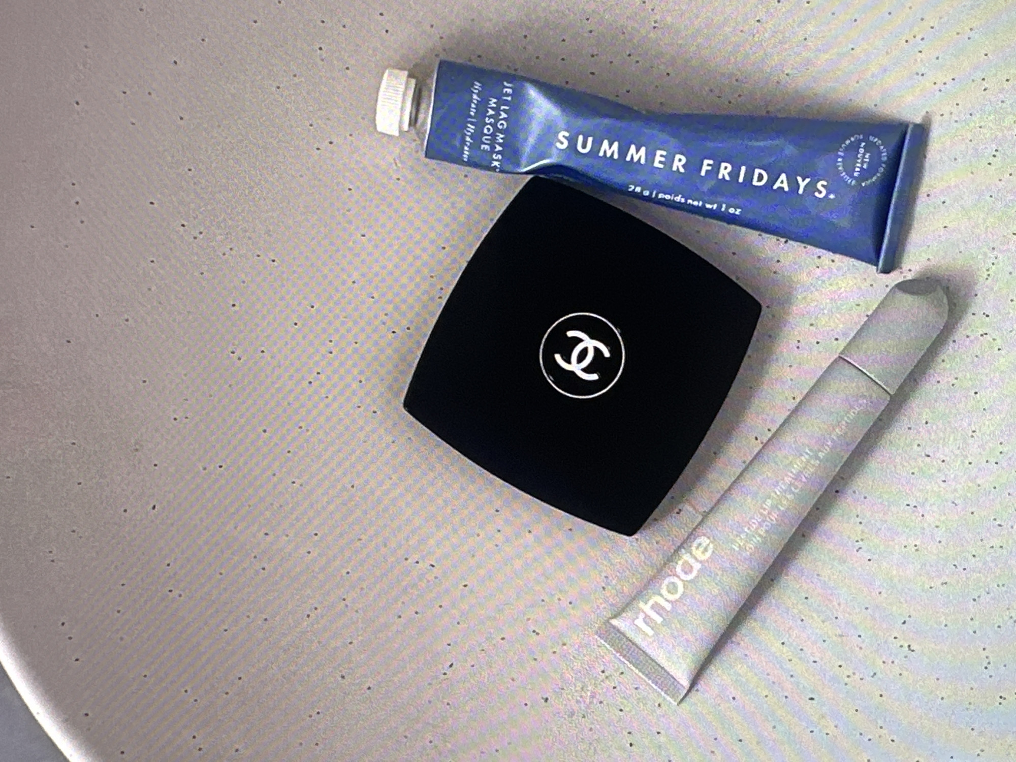 The image displays a cosmetic product arrangement against a textured, light-colored background. There is a black, square compact bearing the Chanel logo, a blue tube labeled "SUMMER FRIDAYS. JET LAG MASK", and a silver tube labeled "rhode. PEPTIDE LIP TREATMENT". The objects are positioned somewhat diagonally on the background, which appears to be a countertop or surface. There are no people visible in the scene. The location is not identifiable from the given image.