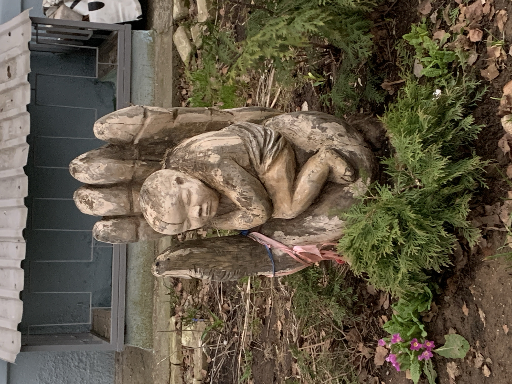 A detailed description of the image, devoid of personal opinions or sentimentality, follows:
The image depicts a sculpture of a human figure resting in the palm of a hand, appearing weathered or aged. The sculpture is in an outdoor setting among plants and soil. It is positioned near the side of a building, with stairs, a metal railing, and siding visible. There is some greenery surrounding the sculpture, including small shrubs and flowering plants with pink blossoms. The location is possibly in the city of Chaikovsky, Russia. The sculpture is tied with a pink and blue ribbon, and looks to be made of wood.
