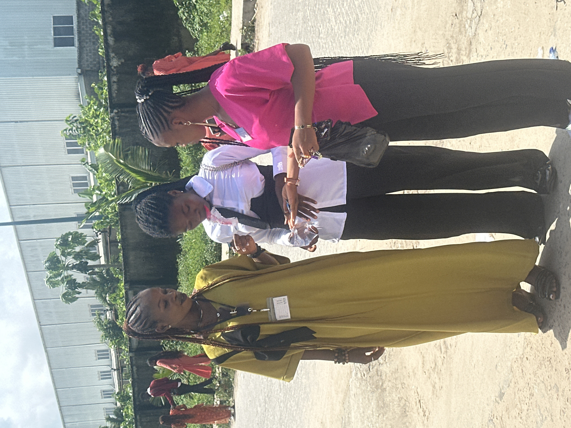 Three young Black women are standing and talking outdoors in what appears to be an industrial or commercial area in Itoga, Nigeria. The woman on the left is wearing a long, mustard-yellow dress and has her hair in braids. She is holding a clear plastic bottle. The woman in the center is wearing a white collared shirt with a dark vest and black pants, her hair also in braids. She is holding a small, clear object. The woman on the right is wearing a bright pink shirt and black pants, her hair in braids. She is holding a black plastic bag and a small ring. In the background, a large, gray, corrugated metal building with multiple windows is visible, suggesting an industrial facility. A concrete wall topped with dark fencing runs along the middle ground, with lush green vegetation behind it. Several other individuals in the background are wearing bright clothing, possibly orange or red, and are walking away from the camera. The ground is a dirt and gravel surface, with some scattered debris. The lighting suggests it is daytime with a bright sky.
