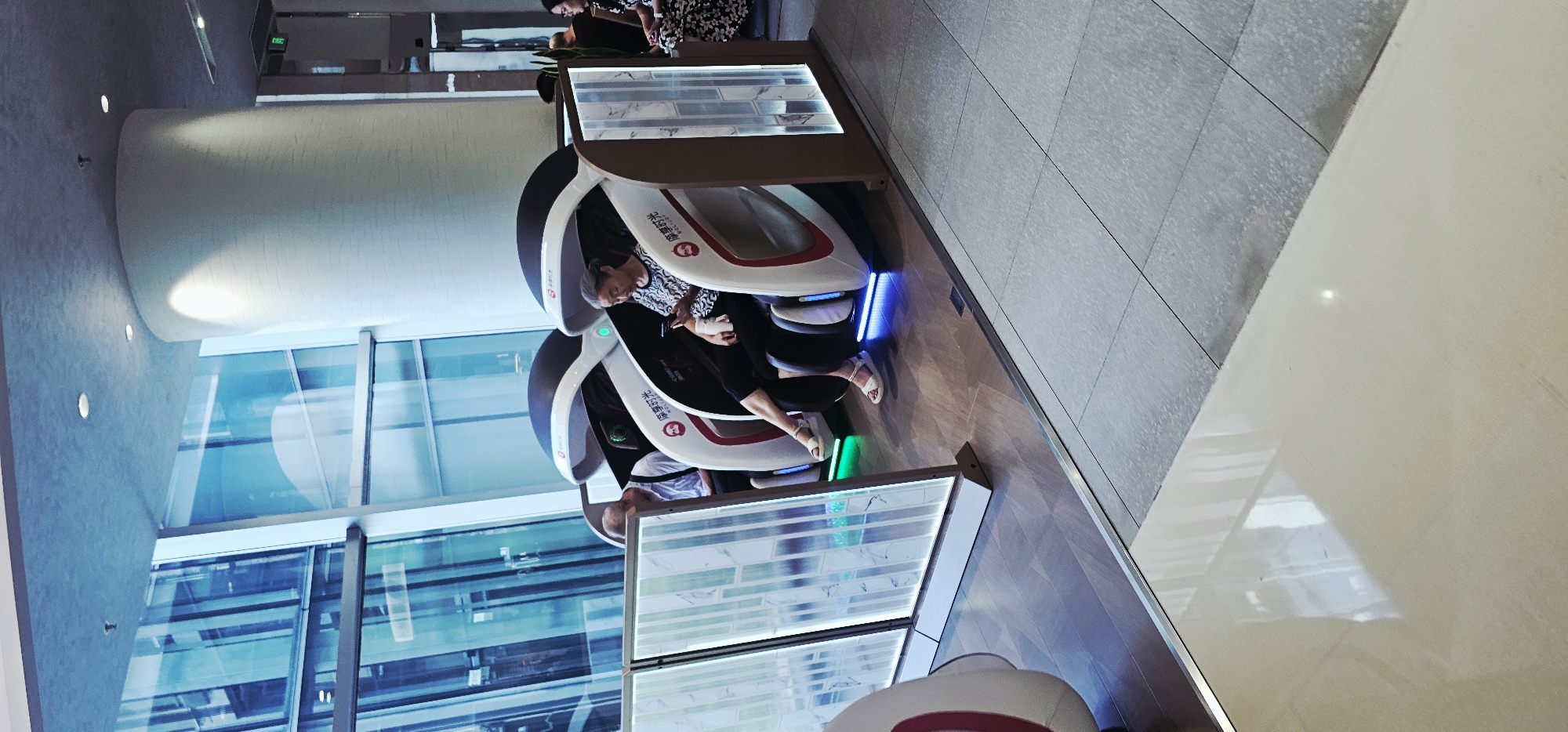 In Jiulongpo District, China, two individuals are seated in what appear to be massage chairs, positioned within a modern interior space. The chairs are futuristic in design, with white and red exteriors and dark canopies. The woman on the right is looking at her phone, while the man on the left is looking down. Both are wearing casual clothing. The chairs are illuminated with colored LED lights at their base, casting a green and blue glow onto the wooden floor. In front of the chairs, a partition with illuminated panels featuring a marble-like pattern separates them from the foreground. Large windows with a blue tint and a grid-like structure are visible in the background, reflecting the interior and possibly the exterior environment. The floor is a mix of polished wood and tiled surfaces. The overall impression is of a waiting area or relaxation zone in a contemporary commercial or public space.