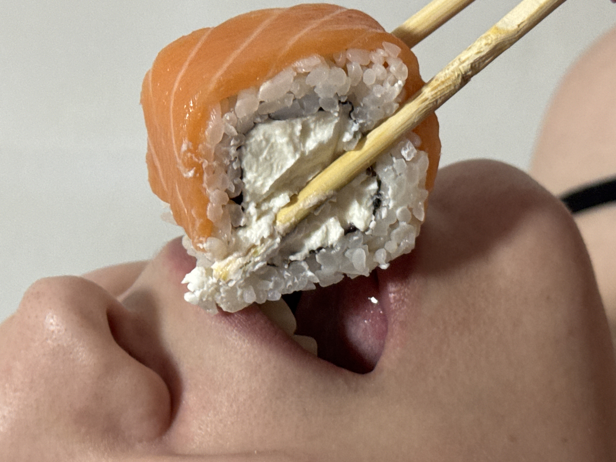A close-up, eye-level shot captures a person in the act of consuming a sushi roll in the city of Chaikovsky, Russia. The individual, primarily identified by the lower portion of their nose, an open mouth with visible tongue and a single tooth, and part of their chin, is presented in profile or near-profile. Skin texture with visible pores is discernible on the nose and surrounding facial areas. A single sushi roll, approximately 4-5 cm in diameter, is held by a pair of light brown, textured wooden chopsticks and is partially inserted into the person's open mouth. The sushi roll features an outer layer of light orange, thinly sliced salmon, encasing a layer of white rice with discernible grains, which in turn surrounds a thick, creamy white filling, characteristic of cream cheese. The background consists of a plain, uniformly light-colored surface, likely an indoor wall, with a thin black strap visible on the lower left side, possibly belonging to the person's attire. Bright, even illumination highlights the textures of the food and skin.