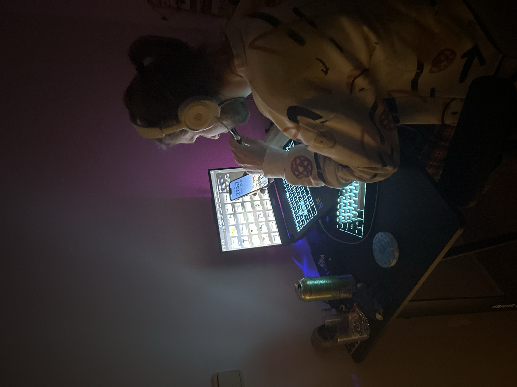 The image shows a person with dark hair in a bun and wearing headphones, seated at a desk. They are looking at a laptop screen displaying a list of items, with a cell phone visible on the screen. A can is present on the desk, along with what appears to be a glass or a cup. The location is not discernible from the image, and there is no indication of Câmara de Lobos, Portugal.