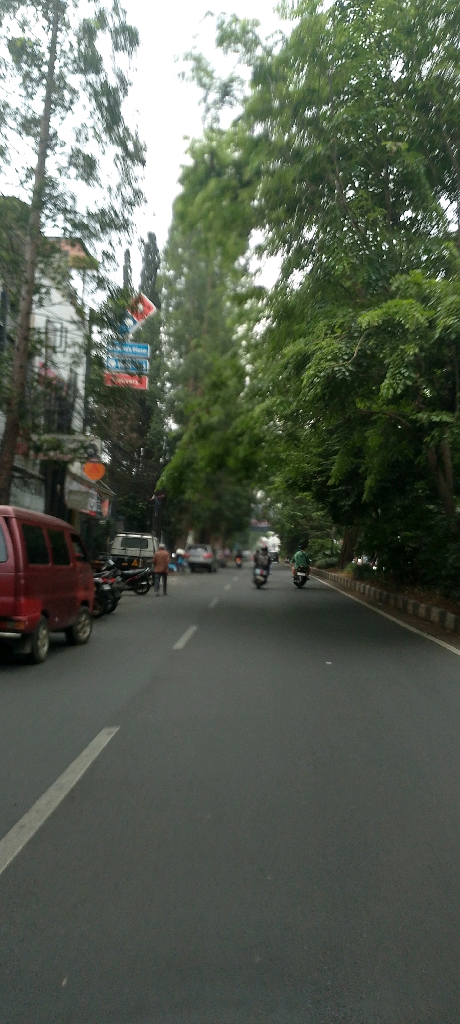 The image depicts a street scene in Bandung City, Indonesia, characterized by lush green trees lining the asphalt road. The perspective is from a moving vehicle, causing a slight blur in the background and some of the objects in the foreground. A red minivan is parked on the left side of the road, with several motorcycles parked behind it. A man in a brown shirt stands near the motorcycles. Further down the road, two people are riding motorcycles, and on the right side, a person in a green shirt is also on a motorcycle. Buildings with signs are visible on the left, and trees create a canopy over the road on the right. The sky is overcast, suggesting a cloudy day.