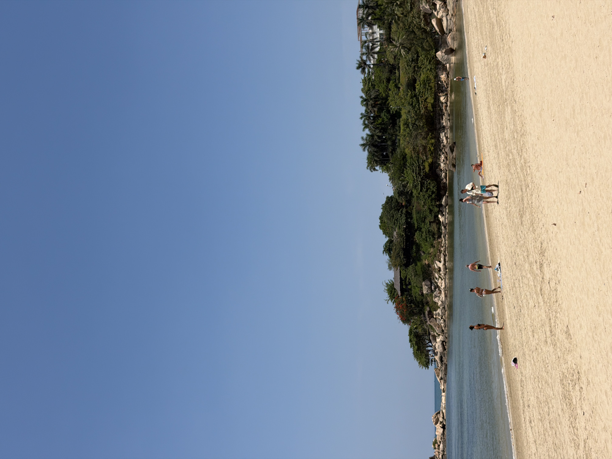 A beach scene on Bac Nha Trang Ward, Vietnam, features a sandy beach meeting calm, light-colored water. Several people are visible on the beach and in the water: A trio of people are standing and appear to be conversing. Further down the beach, three individuals are positioned closely together. In the water near the shoreline, another person stands alone. A section of lush green vegetation lines the edge of the beach. The sky above is a clear, light blue. The image is rotated so the sky dominates the left side of the frame.