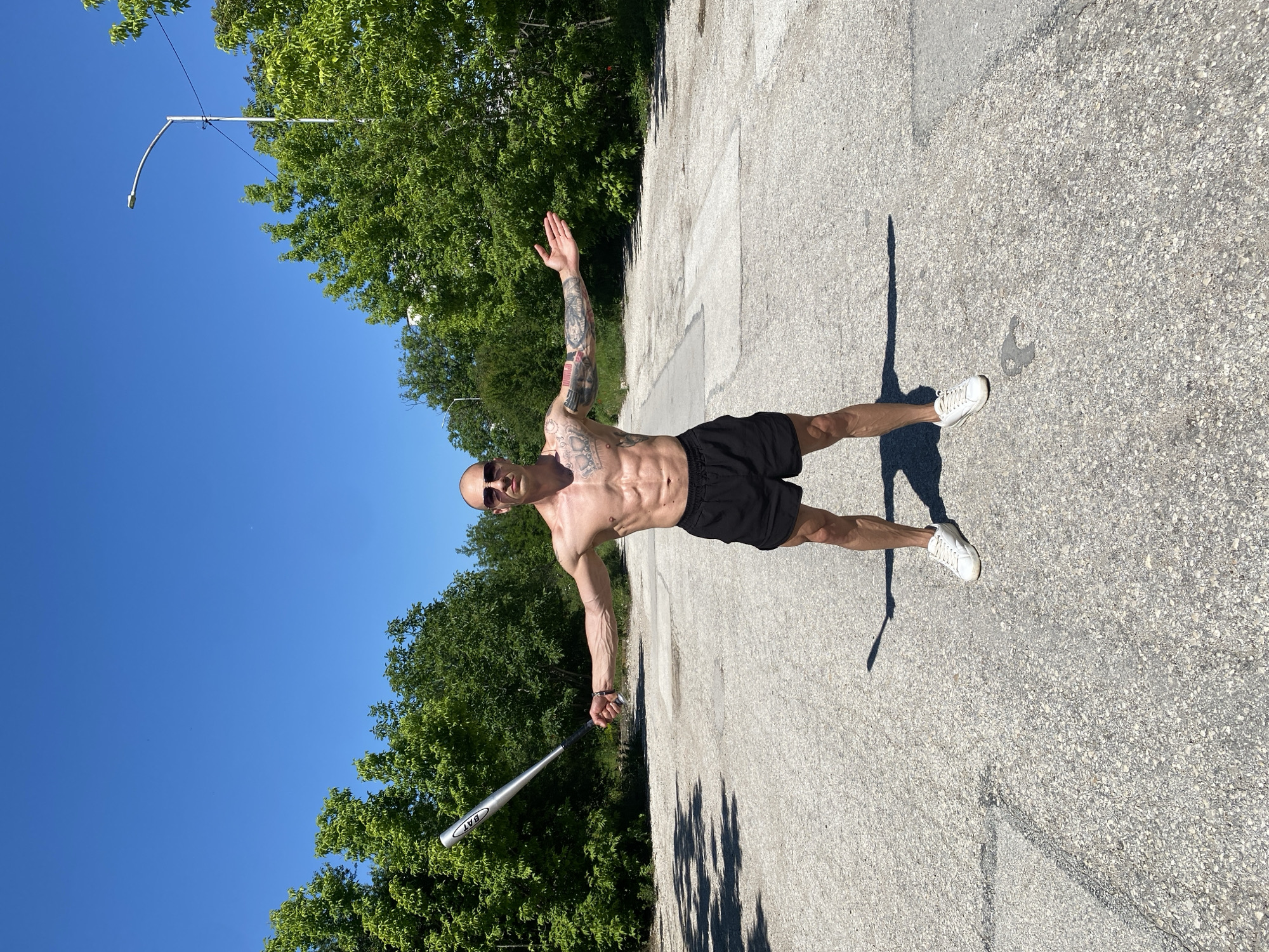 A shirtless man with tattoos, wearing black shorts and white sneakers, stands on an asphalt surface in an outdoor setting. He is bald, wearing sunglasses, and has a silver baseball bat in his left hand. His right arm is raised. The man is casting a shadow. The location appears to be a road or paved area with trees and a street light visible in the background. Based on the image provided, I cannot determine that the location is in Pleven, Bulgaria.