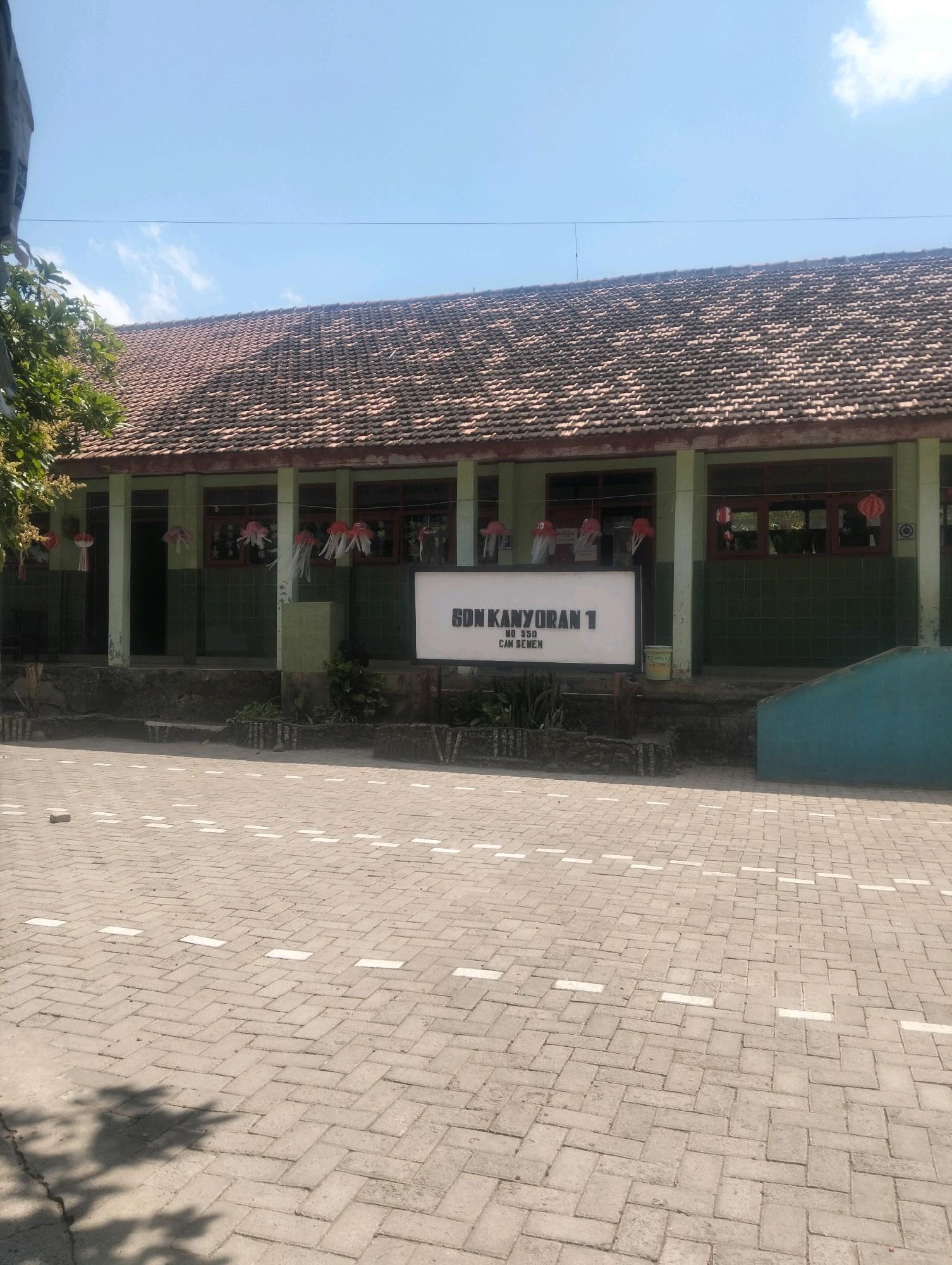 The image depicts the exterior of a school building, identified by a sign as "SDN KANYORAN 1 NO 550 CAM SEMEN" in Kanyoran, Indonesia. The building has a long, tiled roof and a facade with green painted walls and windows. Decorative paper lanterns in red and white are hung along the front of the building. The foreground is a paved courtyard with a herringbone pattern of bricks, marked with white dashed lines. The sky above is clear blue with a few wisps of clouds. There are no people visible in the image.