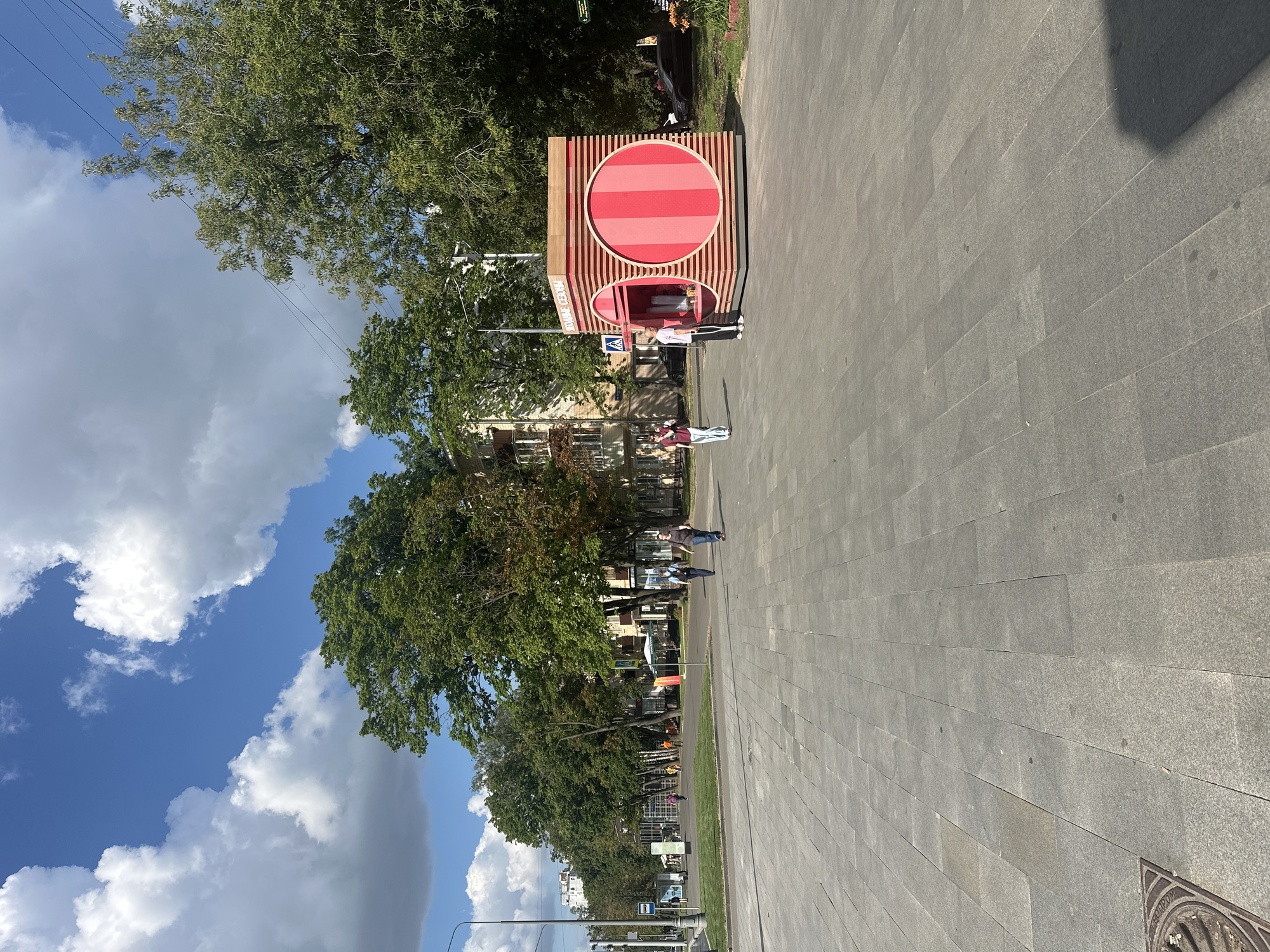 The image captures a sunny day in a city environment, likely Moscow, Russia, as indicated by the Cyrillic text on the kiosk. The scene features a wide, paved pedestrian walkway in the foreground, laid with large, rectangular gray stones. To the right, a modern, cube-shaped kiosk with a vibrant pink and red striped design stands prominently. The kiosk has two circular openings, suggesting it might be a food or beverage vendor. One person, appearing to be a male, is visible near the kiosk, leaning on a crutch and wearing a light-colored shirt and dark pants.
Further in the background, a row of leafy green trees lines a grassy area. Beyond the trees, apartment buildings with visible windows and balconies are present. Several individuals are walking along the sidewalk in the middle ground. A street with cars and a bus stop can be seen on the far left. The sky above is a clear blue with scattered white clouds. The overall impression is of a well-maintained urban public space on a pleasant day.