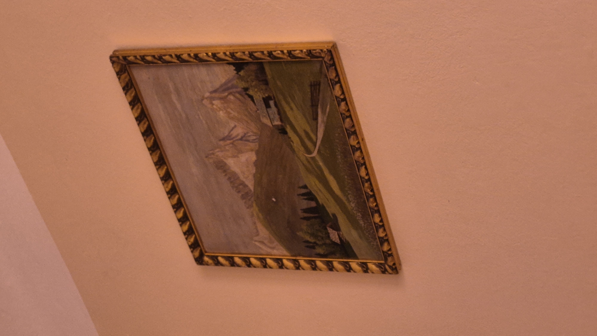 A framed painting is displayed on a peach-colored wall. The painting depicts a mountainous landscape with rolling green hills, scattered trees, and a winding path leading to a small building. The sky is overcast, and the overall impression is serene. The frame of the painting is ornate and golden. The scene is not identifiable as being located in Rzeszów, Poland.