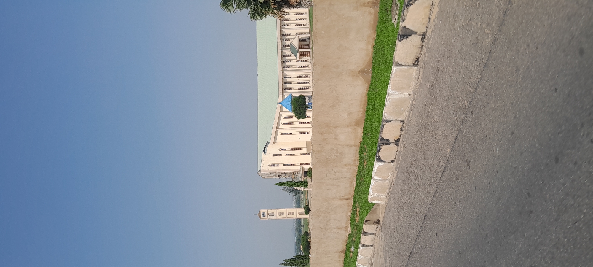 A wide shot captures a large, pale yellow building with a light green roof, possibly a church or a public building, situated in Jalingo, Nigeria. To the left of the main building, a tall, narrow tower with arched windows and a cross on top stands prominently. In front of the building and tower, a rough-textured, beige-colored wall runs horizontally across the frame. Green grass grows in a strip behind the wall, and a palm tree is visible on the right edge of the image. A paved road with a concrete curb forms the foreground, with a portion of a gray asphalt road also visible. The sky is a clear, pale blue. No people or animals are present in the scene.