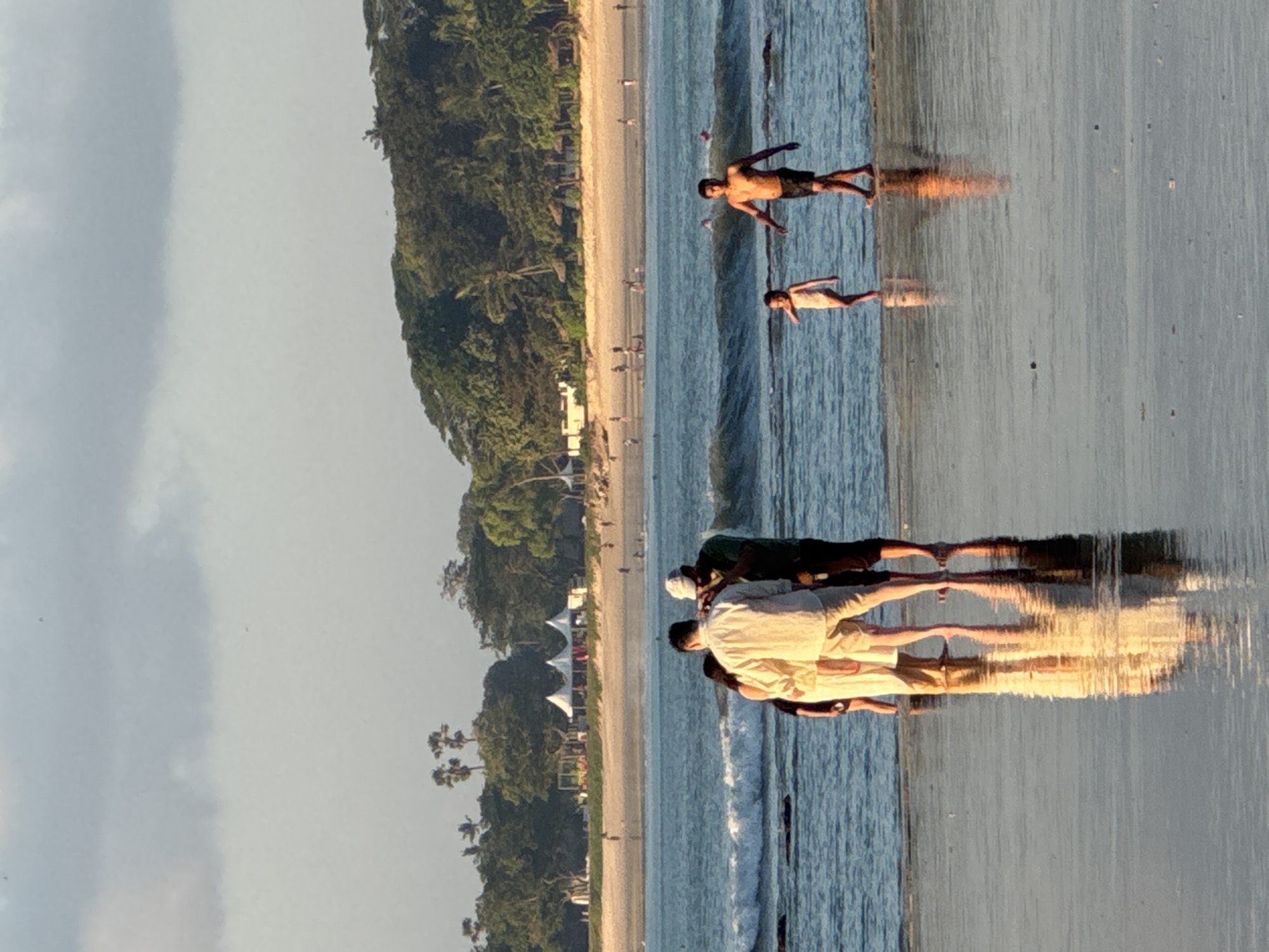 In Kuta Selatan, Indonesia, a group of people are captured on a beach at sunset. In the foreground, three individuals are standing in shallow water, with two of them appearing to interact. A fourth person, a young child, stands a short distance away. Further out in the ocean, a man is walking and a child is in the water. In the background, the beach is populated with more individuals, and palm trees line the shore leading to lush green foliage. The sky is overcast with hints of the setting sun casting a warm glow.