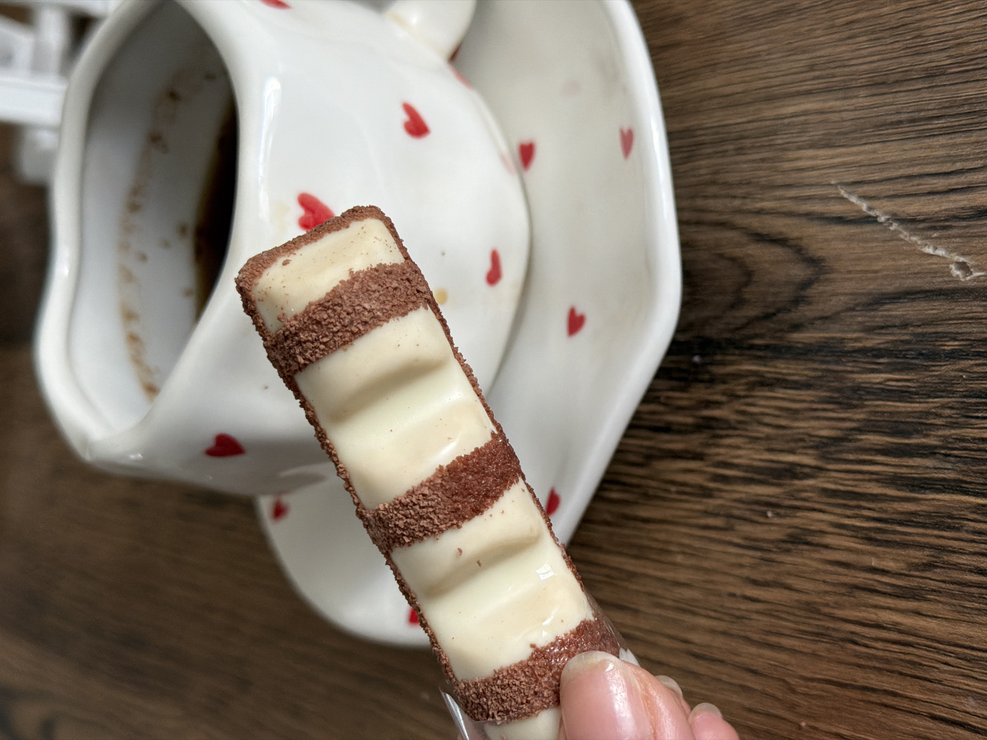 This close-up indoor shot features a hand holding a Kinder Bueno White chocolate bar. The chocolate bar is prominently displayed in the center, characterized by its white chocolate coating, wavy texture, and horizontal stripes of dark chocolate crumbs. The person's fingers are visible, supporting the bar from below, and their nails appear well-kept.
In the background, a white ceramic mug or pitcher with a spout is visible, adorned with small red heart patterns. The inside of the mug contains dark residue, suggesting it recently held a beverage like coffee or hot chocolate. Partially behind the mug, a matching white ceramic saucer or plate with red heart motifs can be seen.
The objects rest on a wooden surface, likely a table or countertop, which shows a distinct grain pattern and some minor scratches or blemishes. The lighting is bright and natural, indicating a daytime setting. The scene evokes a moment of indulgence or a pleasant break with a sweet treat. No text is visible in the image. The location context provided for this image is Perm, Russia.
