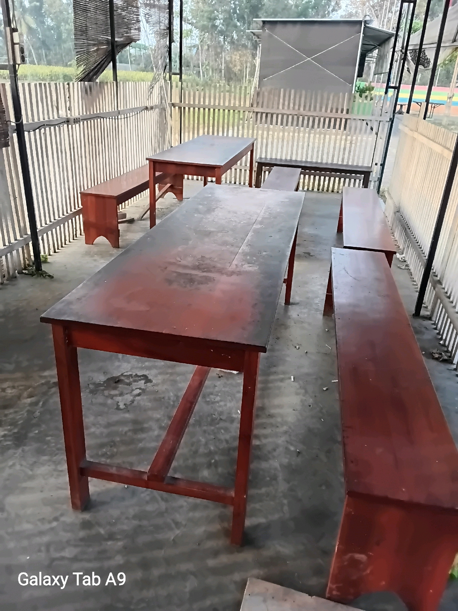 The image depicts an outdoor seating area with multiple red wooden tables and benches. The tables are rectangular, and the benches are long and plain. The ground is paved with concrete and appears somewhat dirty, with scattered debris. A wooden fence runs along the perimeter of the seating area, separating it from a background with trees and vegetation. In the distance, a building or structure with a grey, textured roof is visible. The lighting suggests it is daytime, possibly overcast. The scene is devoid of any people. The overall impression is one of a simple, functional outdoor dining or resting space.