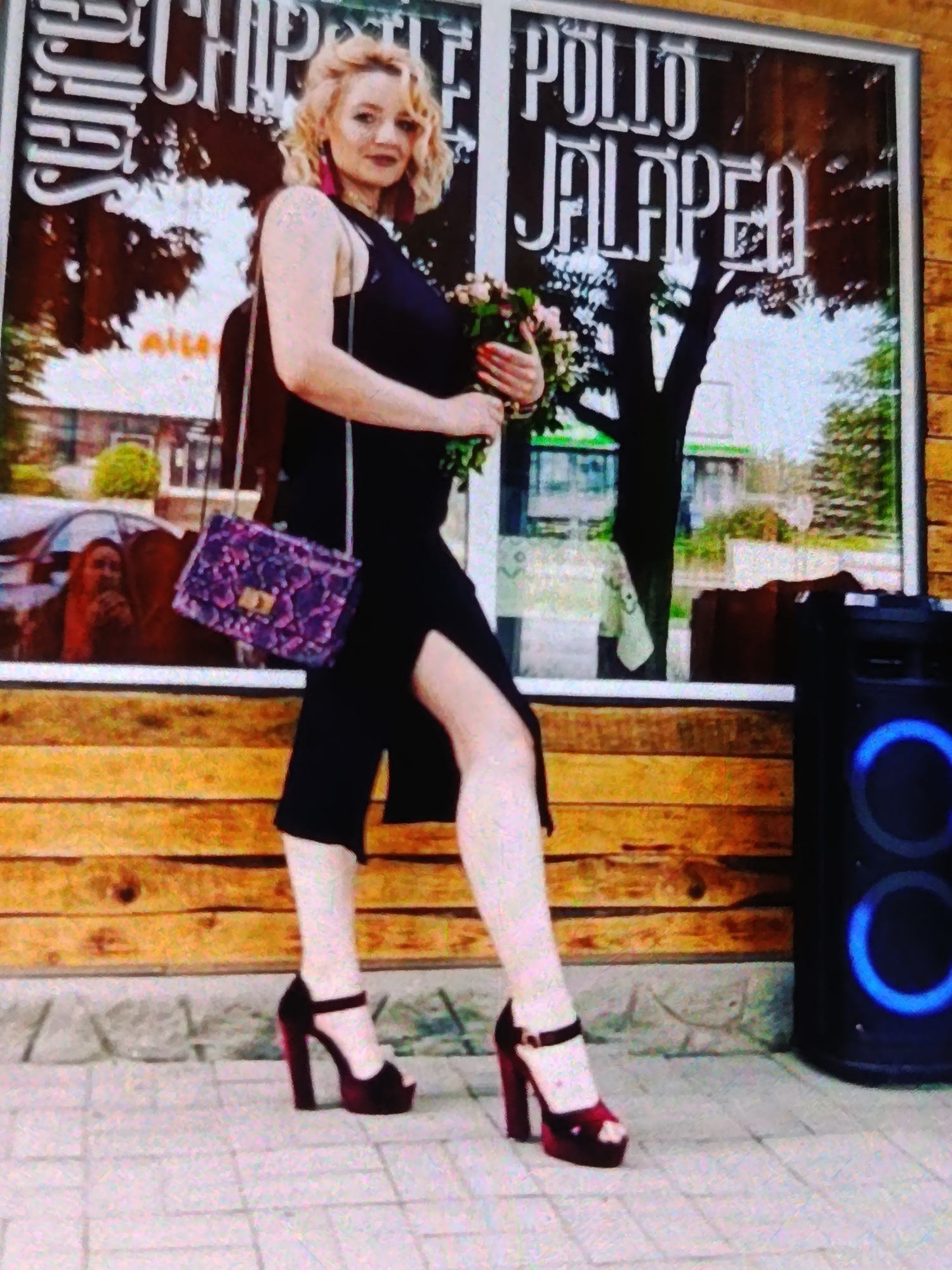 A woman with blonde curly hair and pink earrings stands in front of a storefront. She is wearing a black sleeveless dress with a thigh-high slit and dark red platform heels. In her right hand, she holds a bouquet of light-colored flowers. A purple snakeskin print clutch is slung over her left shoulder. To her right, a black speaker with blue illuminated rings is visible. The storefront behind her has large windows with wooden trim. The window displays text, including "CHIPOTLE", "POLLO", and "JALAPEÑO". The woman appears to be posing for a photo. The scene is outdoors, and the lighting suggests it might be daytime. The paving stones in front of the storefront and the wooden facade give a sense of an urban or commercial setting. The overall impression is one of casual fashion and posing for a photograph.
