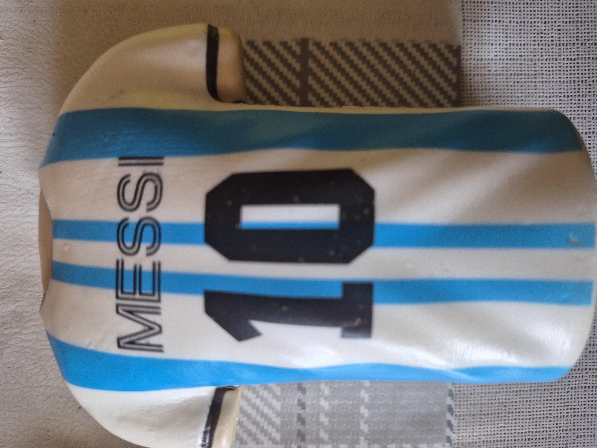 The image displays a celebratory cake designed in the likeness of a football (soccer) jersey, resting horizontally on a patterned surface. The cake features a prominent design of alternating light blue and white horizontal stripes, characteristic of the Argentina national football team's kit.
On the cake, the number "10" is clearly visible in large black font, while the name "MESSI" is written vertically in black capital letters to the left of the number. The cake's shape mimics a jersey, with a slight bulge on either end suggesting the shoulder/sleeve areas, one of which has a black band detail.
The cake is placed on a surface composed of at least two distinct textures. Directly beneath the cake, there's a light gray fabric with a diagonal zigzag or chevron pattern. To the right and partially beneath this pattern, a section of a lighter, possibly white or off-white, finely woven or mesh-like fabric is visible. On the far left and top, a lighter, irregularly textured surface, possibly another fabric or a tabletop, is also present.
The overall impression is that of a themed cake, likely made for a fan of Lionel Messi or to celebrate a football-related event. No people are visible, and the scene is a close-up, static shot of the cake. The lighting is soft and ambient, suggesting an indoor setting. The visible text is "MESSI" and "10".