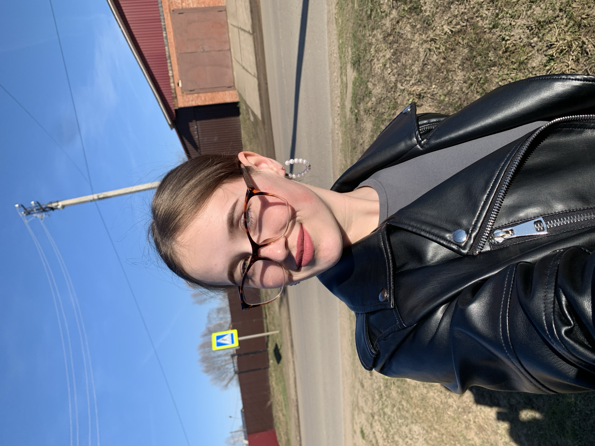 A young woman is captured in a bright, outdoor selfie, rotated to appear on her side. She is looking directly at the camera with a slight, pleasant smile, showcasing light pink lipstick. Her brown hair is partially visible, and she wears glasses with a tortoiseshell frame. A hoop earring adorned with small, light-colored beads can be seen on her right ear. She is dressed in a black, leather-look motorcycle jacket with prominent silver zippers and snaps over a plain grey top.
The setting appears to be a sunny day, likely midday or early afternoon, indicated by the clear blue sky and strong shadows. In the background, to her left, a yellow pedestrian crossing sign with a blue square and a white pedestrian symbol is visible. Further back, a road or path runs alongside grassy terrain, and a utility pole with wires stands against the sky. A building with a reddish roof and a dark, possibly brick, facade is partially visible in the upper background. The overall impression is an everyday outdoor scene in a town or village setting, consistent with a location like Фоки, Russia. There is no discernible text visible in the image.