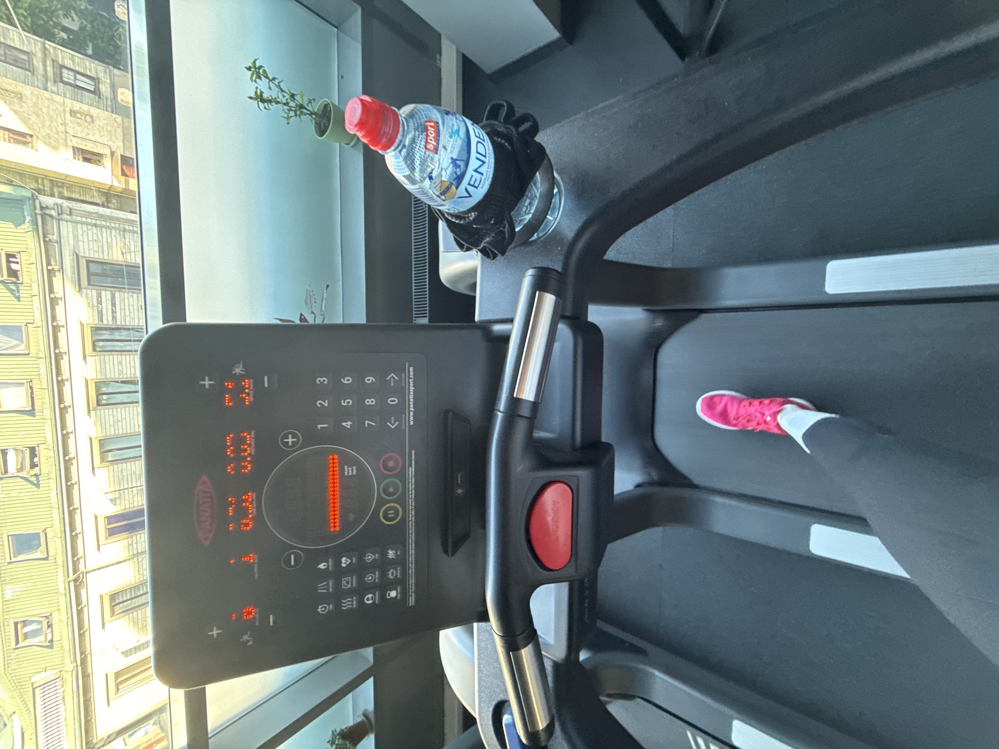 A first-person perspective shot shows a person's legs and feet on a treadmill in a gym. The person is wearing black leggings and bright pink athletic shoes. The treadmill console displays various metrics, including speed, distance, and heart rate, all illuminated in red LED lights. A bottle of "VENDE" brand water with a red cap is secured in a cup holder on the treadmill. In the background, through a window, buildings with green roofs and numerous windows are visible, suggesting an urban setting. The architecture of the buildings implies the location is Riga, Latvia.