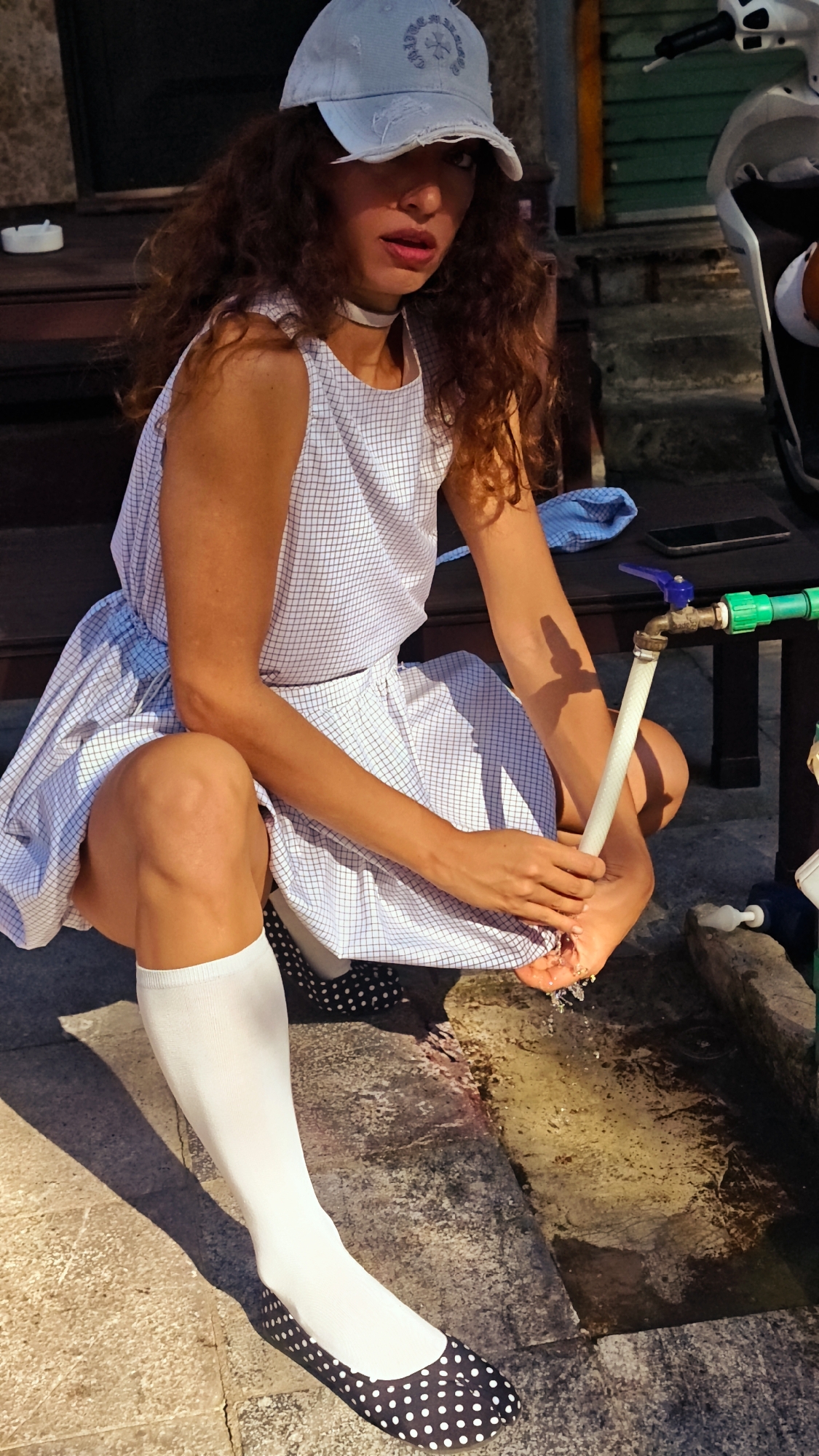 In Bắc Hà, Vietnam, a woman squats outdoors, washing her hands under a faucet. She wears a light-colored, plaid dress with a matching belt, white knee-high socks, and dark flats with white polka dots. A distressed white cap sits on her head. The faucet is connected to a green pipe and empties into a stone trough. Behind her, a dark wooden bench holds a cell phone and a blue cloth. A motorbike is visible on the right edge of the frame.