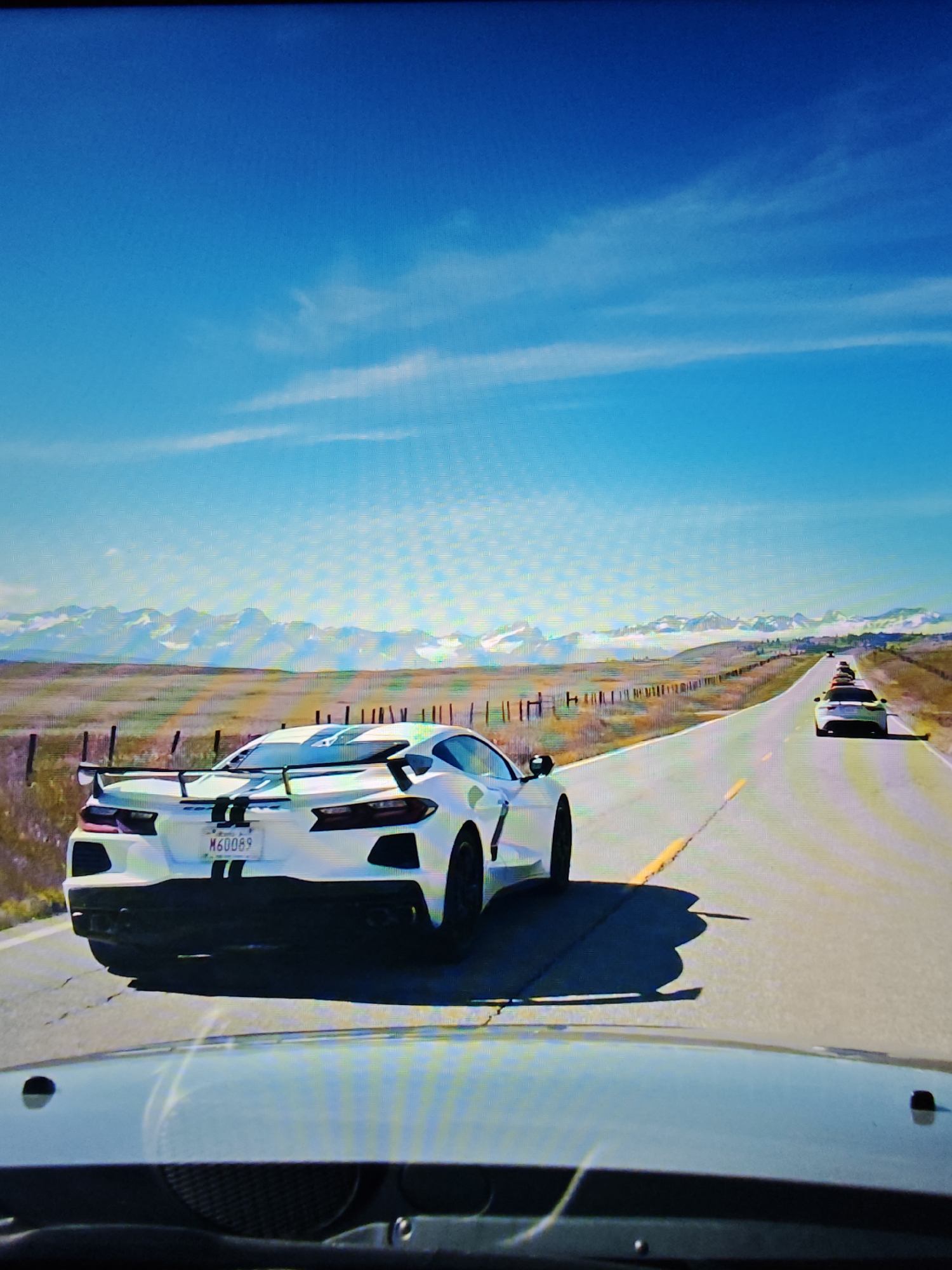 The image captures a convoy of white sports cars driving on a road with a mountain range in the background. The lead car, a white Corvette with racing stripes and a spoiler, is in the foreground. The license plate reads "M60069". The road is paved with yellow lines indicating lanes. The landscape is dry and sparse, with a wooden fence running alongside the road. The sky is a clear blue with a few wispy clouds. This scene is set in the Canadian Rockies, a mountain range that forms the southeastern boundary of Alberta, Canada. The image does not provide specific details about the city of Calgary, Canada, other than its proximity to the Rocky Mountains.