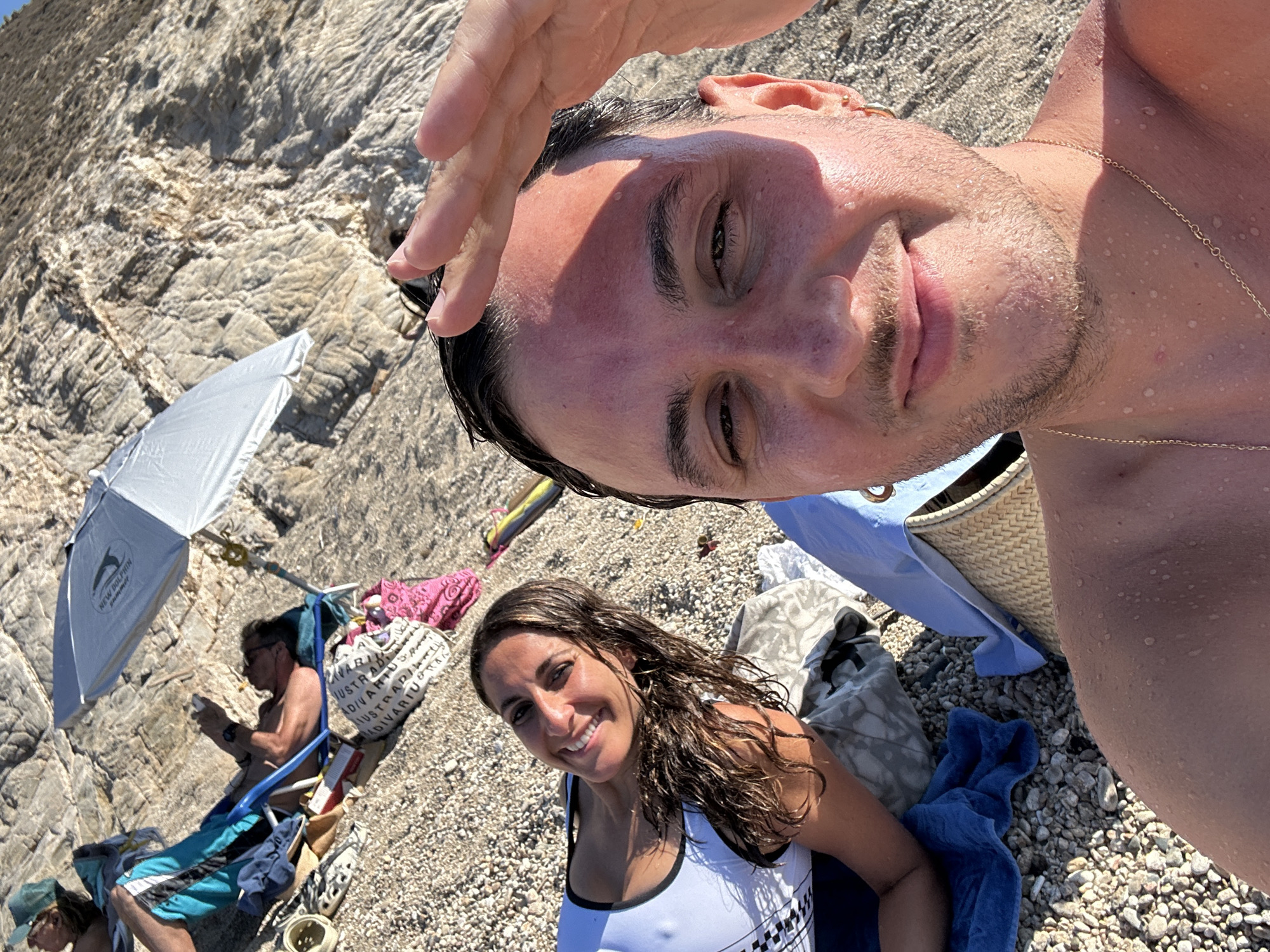 In Ano Mera, Greece, two individuals are enjoying a day at the beach. In the foreground, a man, partially visible, is taking a selfie, holding his hand up to his forehead. He is wearing a gold chain. To his left, a woman is lying down, smiling at the camera. She has long, wet, brown hair and is wearing a white tank top with black trim. Behind them, a rocky cliff face dominates the background. Several beach items are scattered on the pebble-strewn ground, including a beach umbrella, a blue towel, and a woven beach bag. In the middle ground, another person is reclining on a blue beach chair, partially shaded by the umbrella, and appears to be looking at their phone.