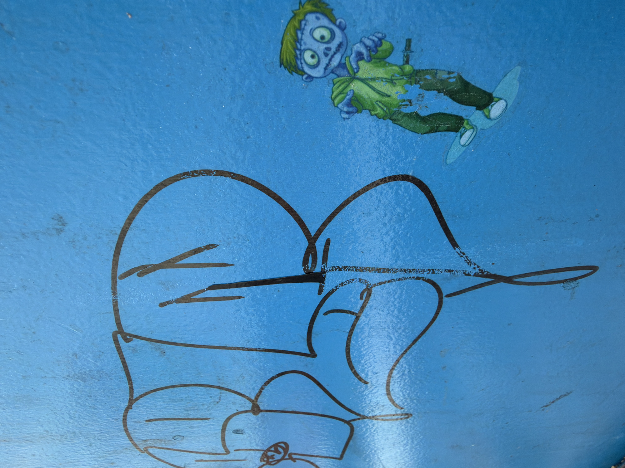 The image depicts a section of a light blue surface, possibly metal or painted wood, bearing two distinct visual elements.
In the upper portion is a partially deteriorated sticker of a cartoon zombie. The zombie has pale blue skin, green hair, and wears a green shirt and dark pants. It appears to be riding a light blue skateboard.
Below the zombie sticker is a large, crudely drawn cartoon character in brown marker. It appears to have large eyes and ears and be wearing a hat. The marker lines are thick and show some variation in pressure.
The background is a solid light blue color with variations in texture, suggesting it may be painted or coated. The presence of both a sticker and marker drawing suggests this may be a surface accessible to the public.
The image does not provide enough information to confirm if this is located in Chaikovsky, Russia.
