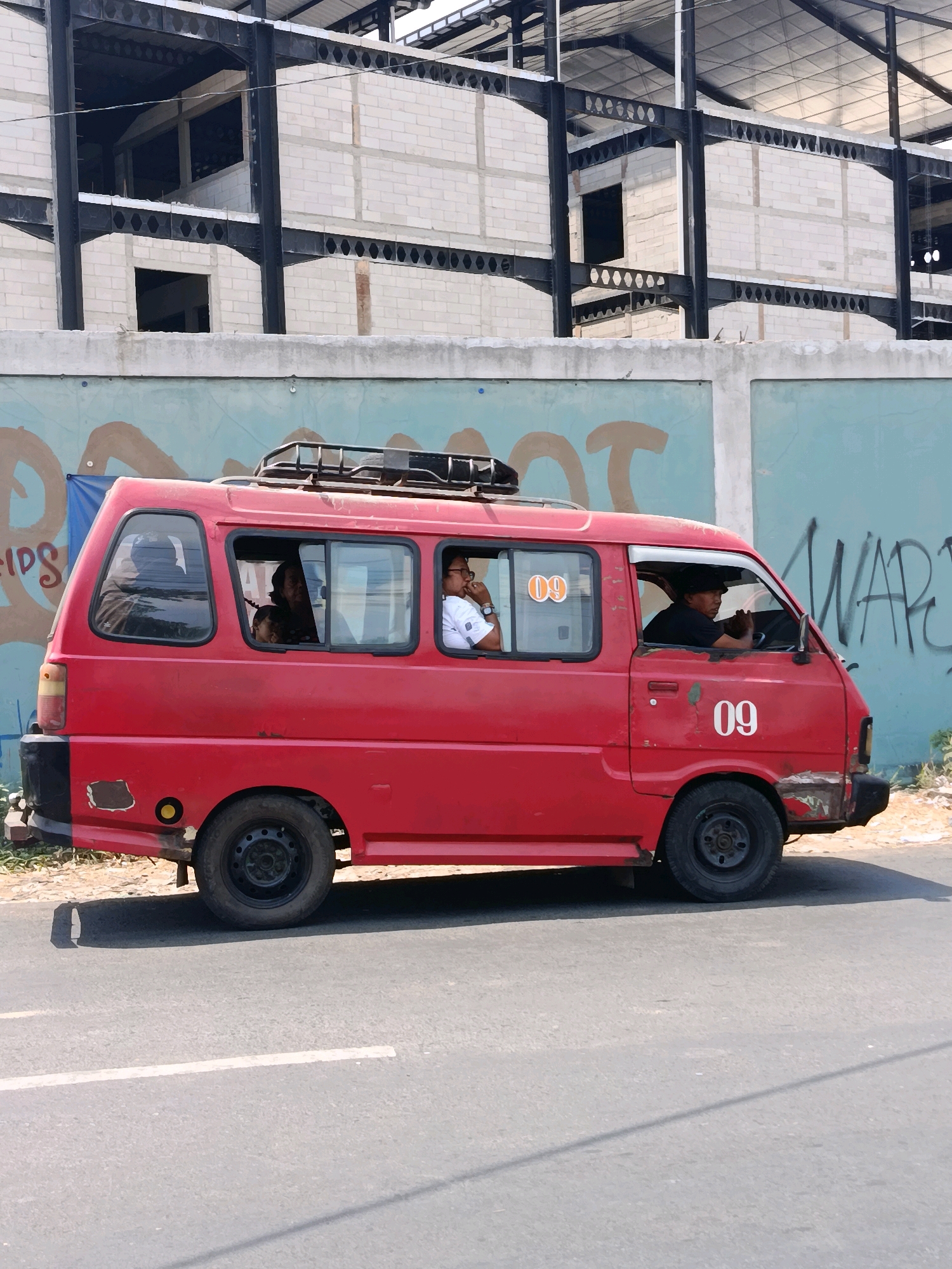 A bright red van, possibly a public transport vehicle, is captured in a side profile shot on a road in Babelan, Indonesia. The van has the number "09" displayed prominently on its side and also on one of its windows. The van's windows are partially tinted, revealing silhouettes or faces of passengers inside. The driver, wearing a black cap, is visible behind the steering wheel. A rack is mounted on the roof of the van.
The background features a partially constructed building with exposed steel beams and concrete blocks, suggesting an ongoing construction project. A blue wall with graffiti-like markings is visible behind the van, contributing to an urban, possibly industrial, setting. The road is paved and appears to be a typical street in the area. The lighting suggests it is daytime.