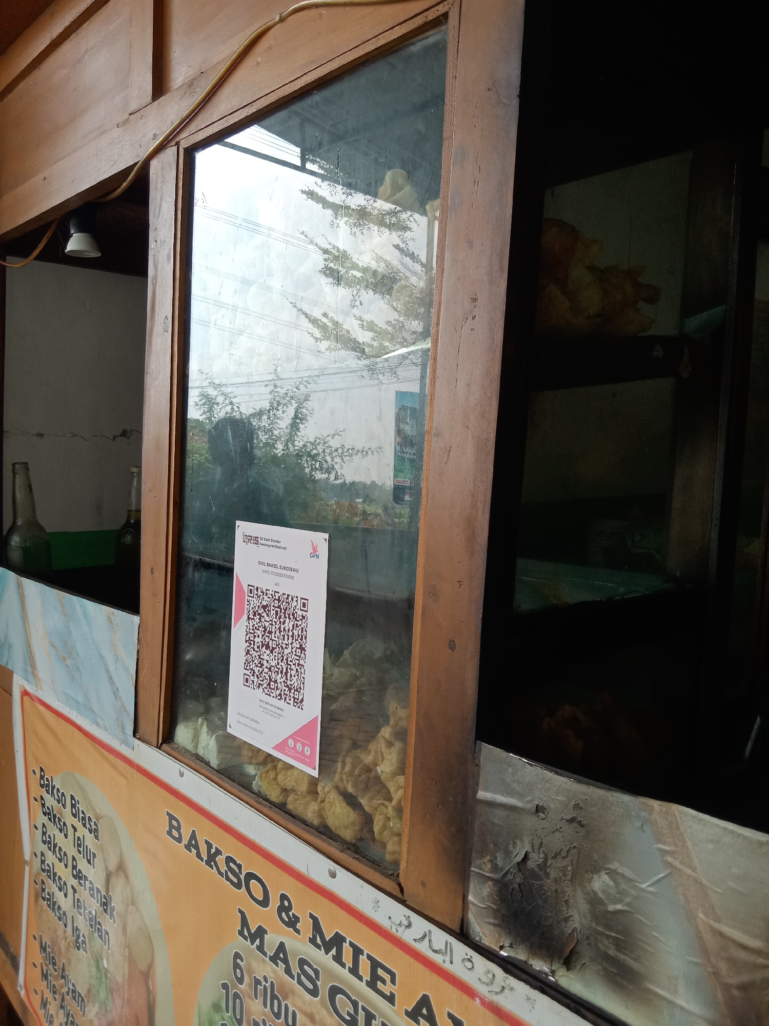 The image shows a food stall in Sugihwaras, Indonesia, with a variety of dishes displayed. The stall appears to be made of wood and glass, with a sign advertising "Bakso & Mie Ayam". There is a QR code displayed for payment. The food items visible include bakso (meatballs) and mie ayam (chicken noodles), along with other fried items. The scene is set outdoors, with trees and a cloudy sky visible in the background.