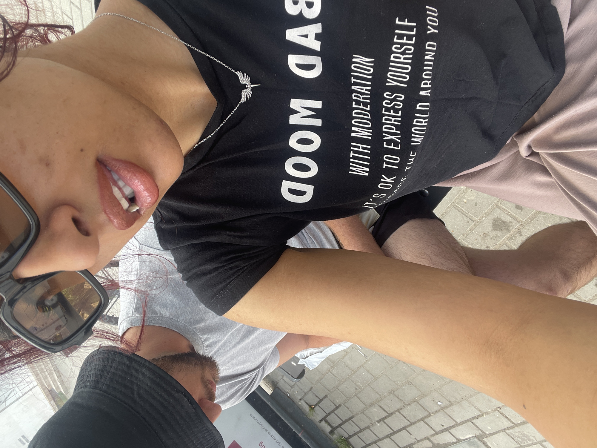 An unidentified outdoor urban location features two individuals. In the foreground, partially visible, is a person with light brown skin, wearing large black sunglasses and a black t-shirt. Dark reddish hair strands are visible near the ear. The t-shirt displays white text: "BAD MOOD" (reversed), "WITH MODERATION", "IT'S OK TO EXPRESS YOURSELF", and partially visible "THE WORLD AROUND YOU". A silver chain necklace with a small, winged pendant adorned with clear stones is worn around the neck. The person's lips are painted with a light reddish-brown lipstick, and teeth are slightly visible. A light brown/mauve garment covers the lower body. The person's right forearm is visible, resting on a knee.
Behind and to the left, a second individual, appearing male, is partially visible. This person wears a black bucket hat, a grey t-shirt, and has a beard. The individual's head is tilted downwards.
The ground is paved with light-colored square or rectangular pavers. Discarded cigarette butts and a small piece of white paper are visible on the ground near the second individual. A dark surface, possibly part of a vehicle or structure, with faint, unidentifiable white and red text is in the background. Bright daylight illuminates the scene, casting shadows.