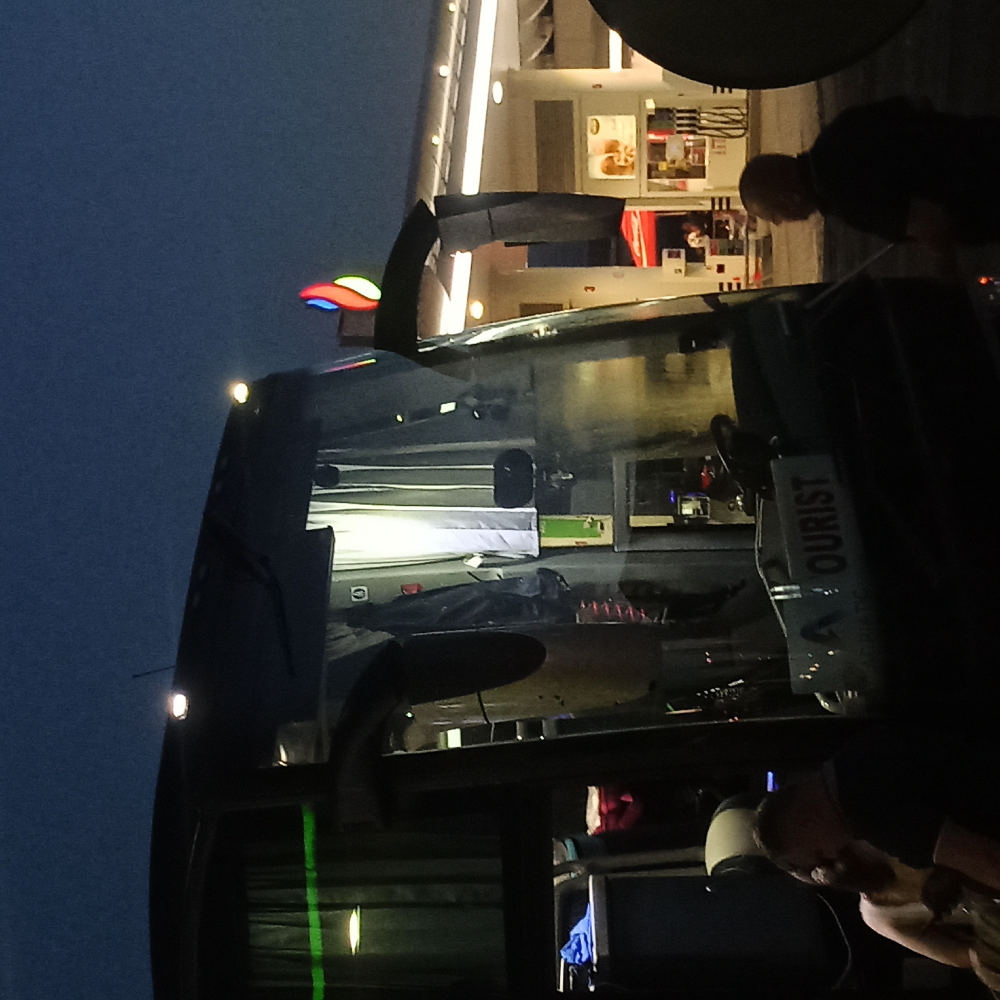 The image captures a dimly lit scene at dusk or night, likely at a gas station, given the presence of fuel pumps and a canopy structure in the background. In the foreground, the side of a dark-colored bus is prominently featured, with its headlights and interior lights illuminated. The text "TOURIST" is visible on a sign on the bus. A person, appearing to be a man wearing a black shirt, is seen from the back on the right side of the frame, facing away from the bus and towards the gas station. Another individual, a woman wearing a light-colored top, is partially visible to the left of the man. The gas station features illuminated signage and structures typical of such an establishment. The sky is dark blue, indicating twilight or nighttime. The location is identified as Adjud, Romania.